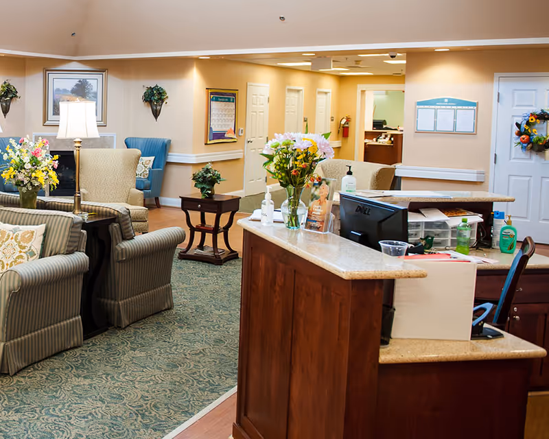A cozy senior living facility common area with a wooden reception desk in the foreground, decorated with flowers and hand sanitizer. Behind the desk, there are comfortable armchairs and sofas arranged around a small table with a lamp and floral arrangements. The walls are painted beige with framed artwork and a bulletin board. The carpet is patterned in green tones, and there are doors and a hallway visible in the background.