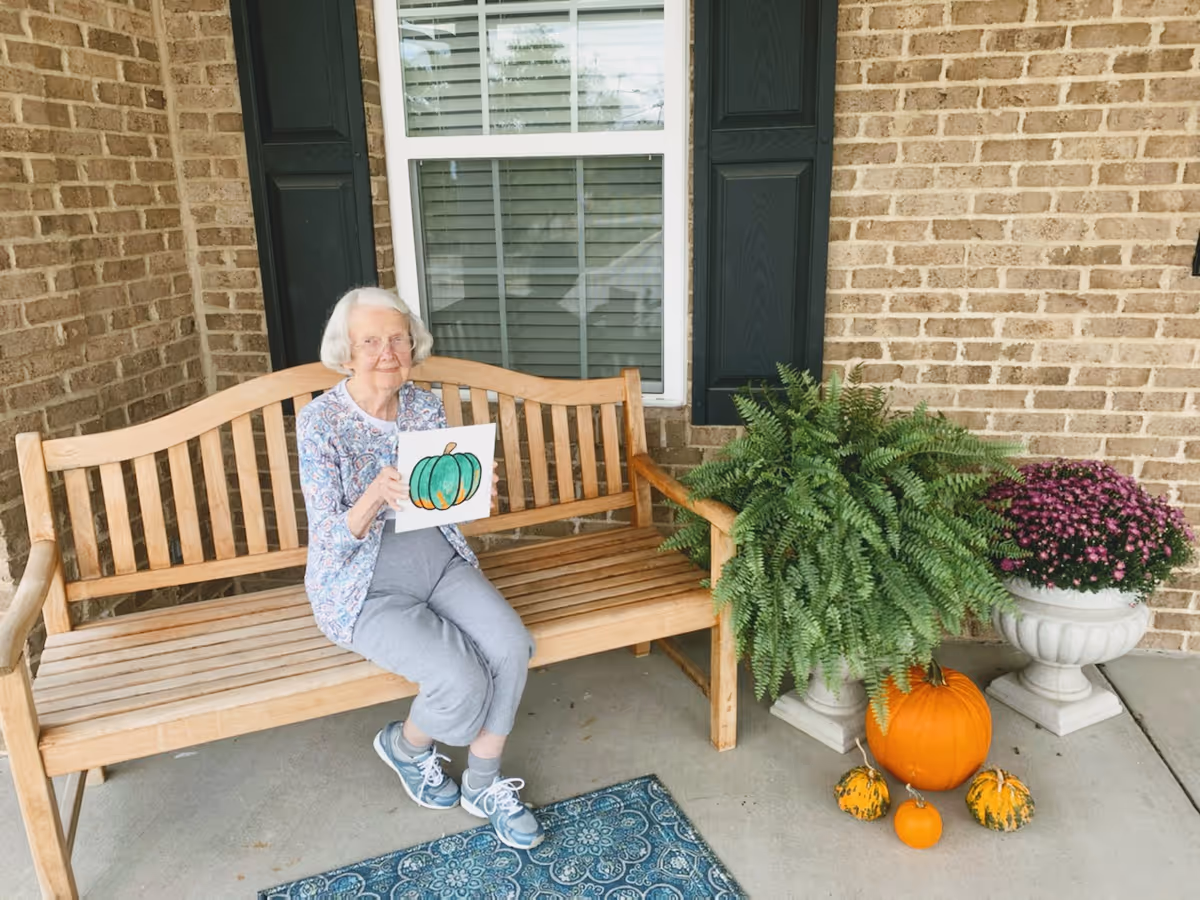 An elderly woman with white hair and glasses sits on a wooden bench outside a brick building. She is holding a drawing of a green and orange pumpkin. Next to the bench are a large green fern, a pot of purple flowers, and several pumpkins arranged on the ground. A blue patterned rug is on the concrete floor in front of the bench.