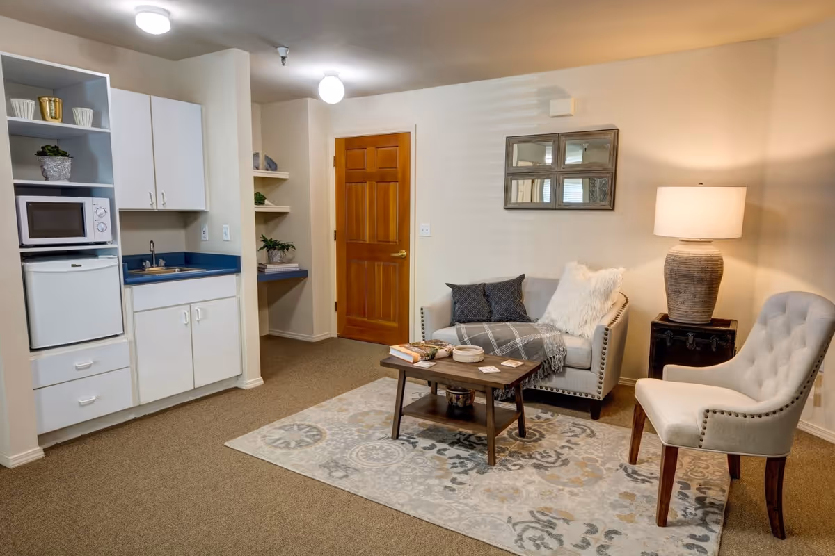 A cozy living area in a senior living facility featuring a small kitchenette with a microwave, mini fridge, and sink on the left. The room has a beige carpet and walls, a wooden door, a small loveseat with pillows and a throw blanket, a wooden coffee table with books and decor, a cushioned armchair, a side table with a large lamp, and a decorative wall mirror.