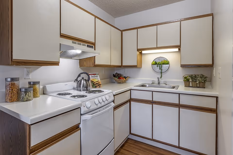 A compact kitchen with white countertops and beige cabinets with wooden trim. The kitchen features a white electric stove with a kettle on top, a range hood with a light, a stainless steel sink, and several decorative items including jars with pasta, a cookbook, a basket of fruit, and small potted plants.