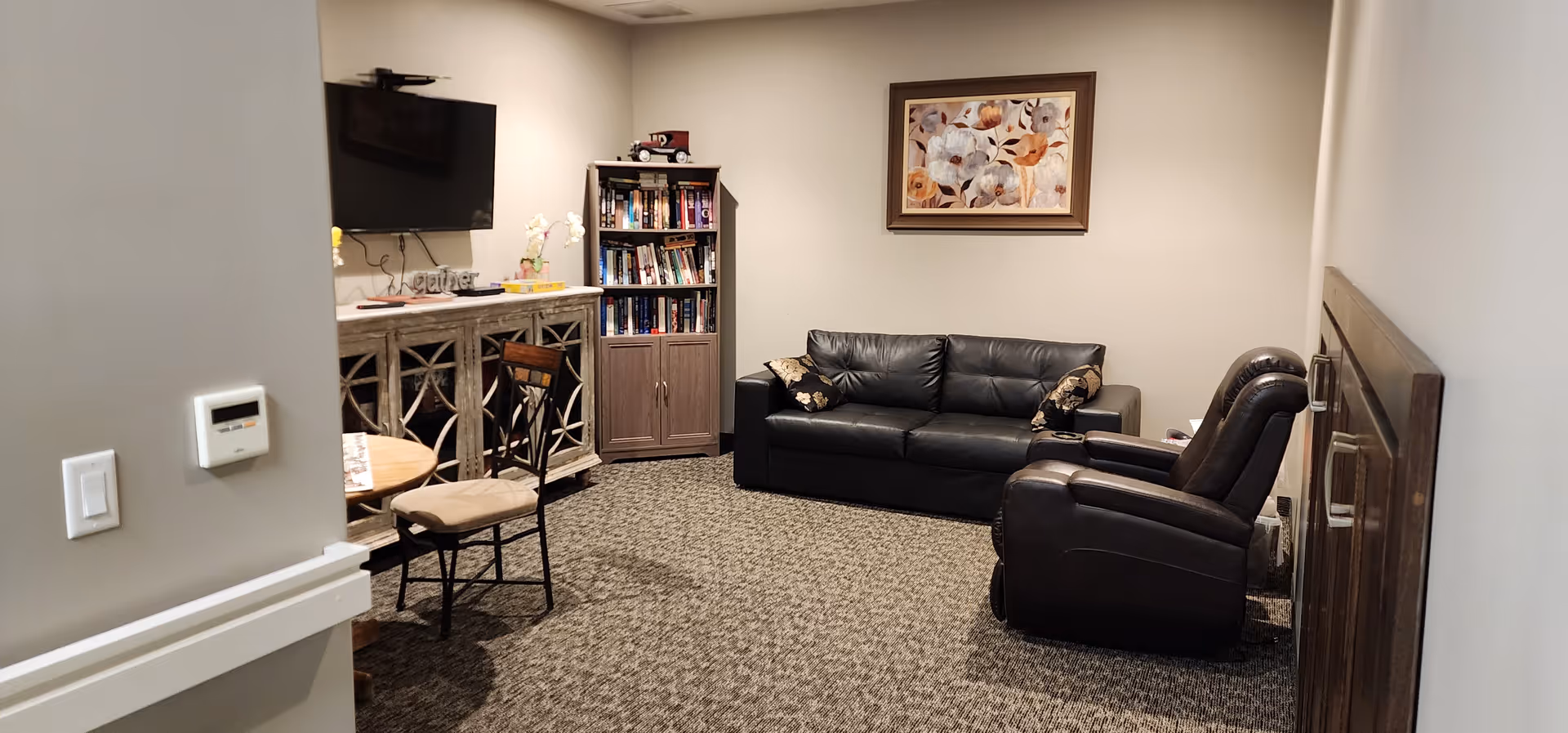 A cozy living room with a black leather sofa and a matching recliner chair. There is a wooden cabinet with a TV mounted above it on the wall. A bookshelf filled with books stands in the corner. A round wooden table with chairs is partially visible. The walls are light-colored, and a framed floral painting hangs above the sofa. The floor is carpeted with a patterned design.