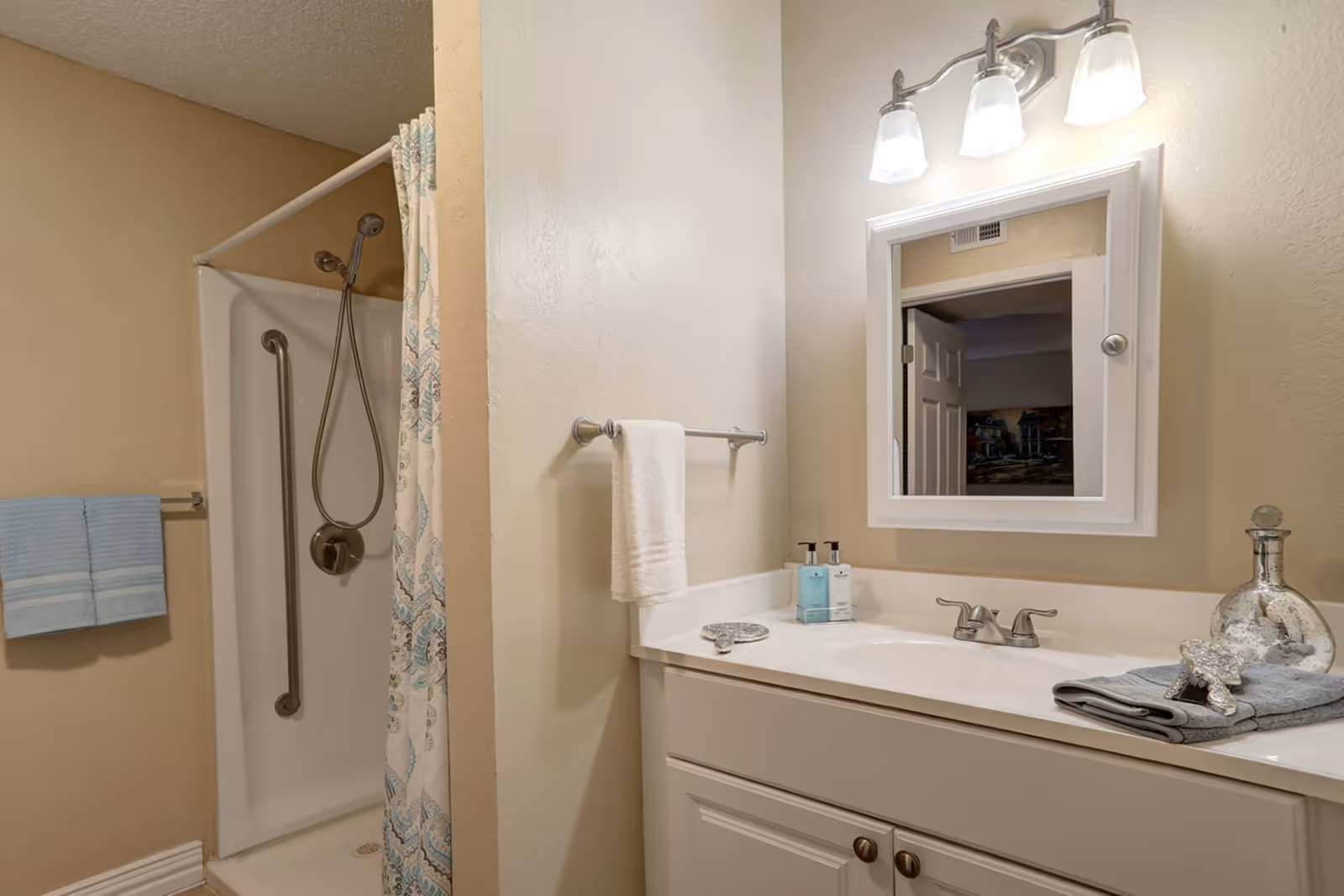 A bathroom with a white vanity and sink, a mirror with three lights above it, a towel rack with a white towel, a shower with a handheld showerhead and a patterned shower curtain, and a blue towel hanging on a wall-mounted towel rack.