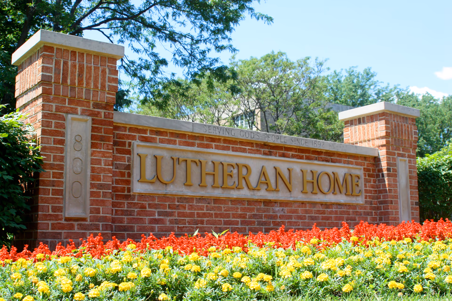 A brick sign with the words 'LUTHERAN HOME' in large gold letters, surrounded by red and yellow flowers in front. The sign also has the number 800 and the inscription 'SERVING GOD'S PEOPLE SINCE 1892' on the top edge. Trees and a building are visible in the background under a clear blue sky.