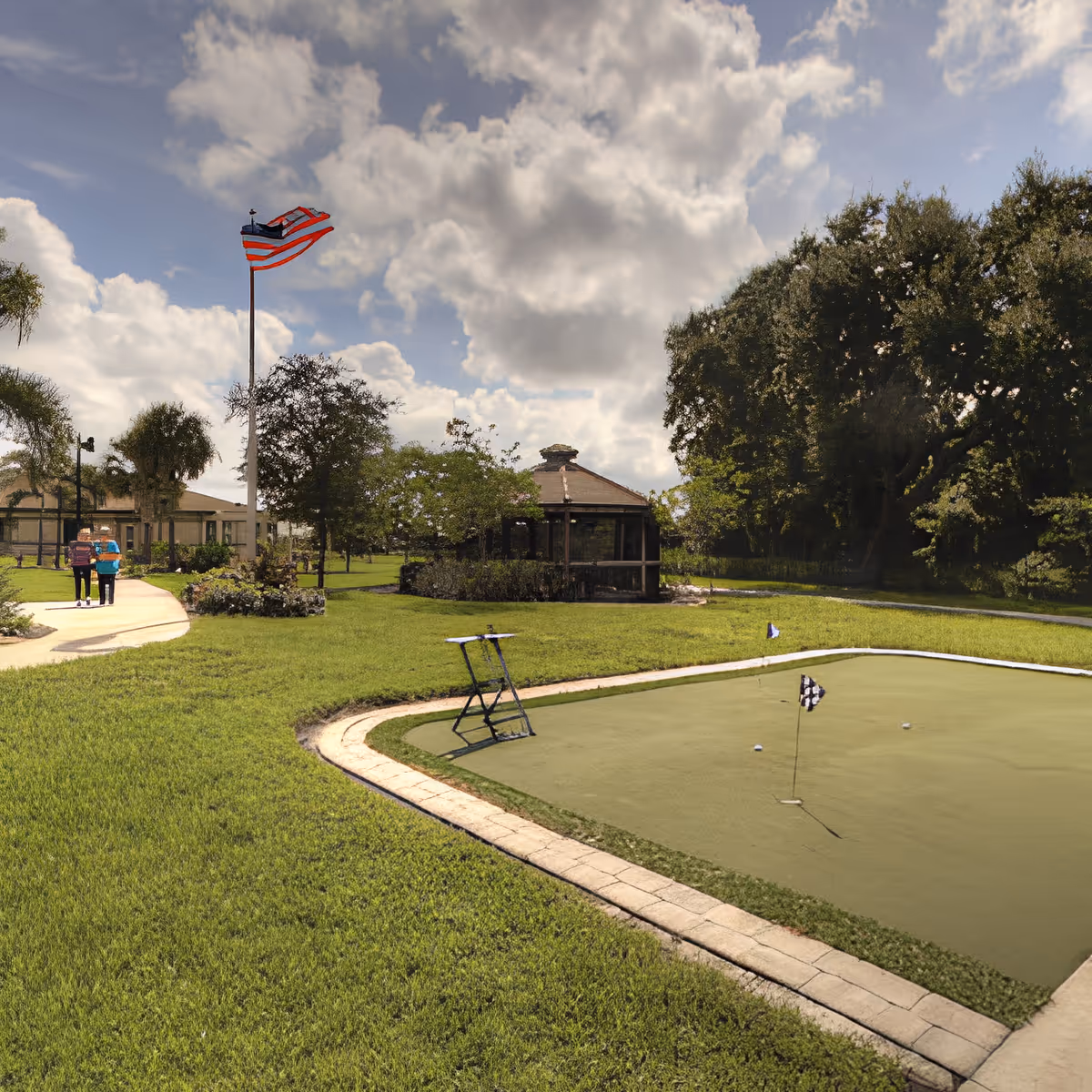 Outdoor scene at Inspired Living Tampa featuring a small putting green with golf flags, a paved walking path, an American flag on a tall flagpole, trees, and two people walking in the distance near a building and a gazebo.