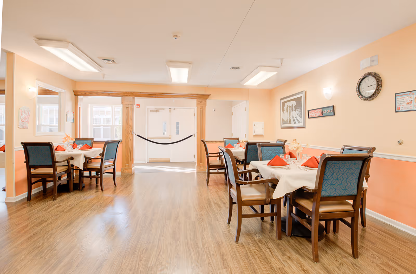 A bright dining room in a senior living facility with wooden floors, peach-colored walls, and several tables set with white tablecloths, red napkins, and glassware. There are wooden chairs with blue patterned cushions around the tables. The room has overhead fluorescent lighting, wall decorations including a clock and framed pictures, and a double door at the far end.