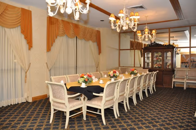 A formal dining room with a long rectangular table covered with a yellow tablecloth and a black runner. The table is surrounded by upholstered chairs and decorated with floral centerpieces and teacups. The room features patterned carpet, large windows with sheer curtains and gold valances, ornate chandeliers, and a wooden china cabinet with glass doors.