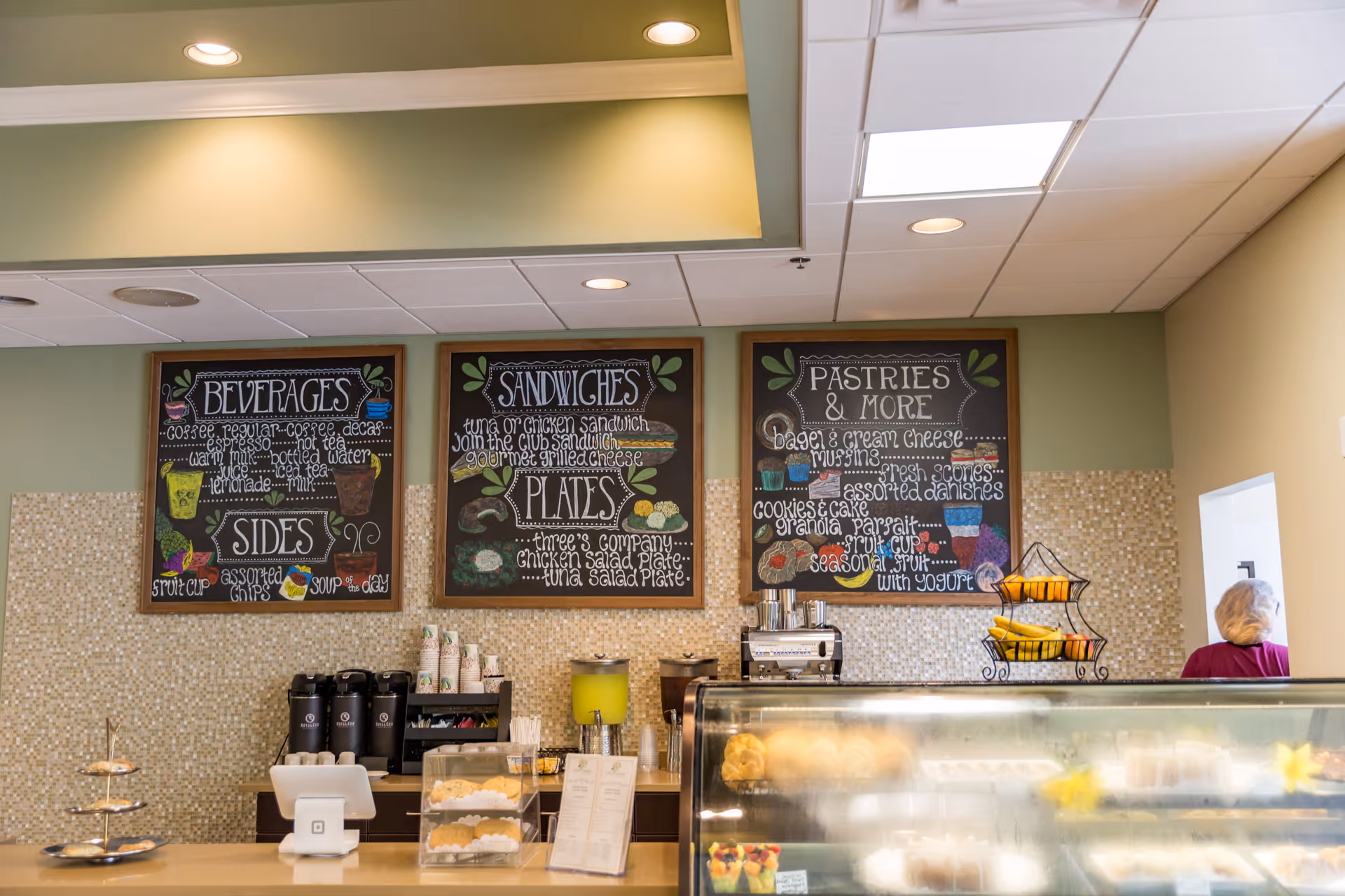 Cafe counter with chalkboard menus reading Beverages, Sandwiches, Pastries & More above a pastry display and coffee station.