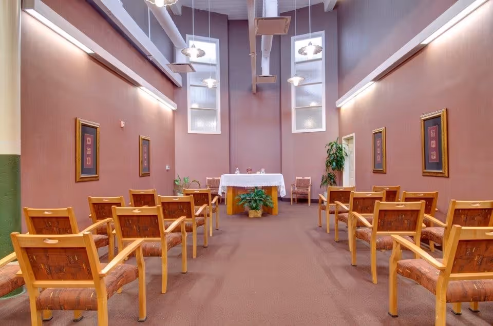 Interior view of a small chapel or meditation room with rows of wooden chairs facing an altar covered with a white cloth. The room has high ceilings with exposed ducts, tall windows, framed artwork on the walls, and potted plants near the altar.