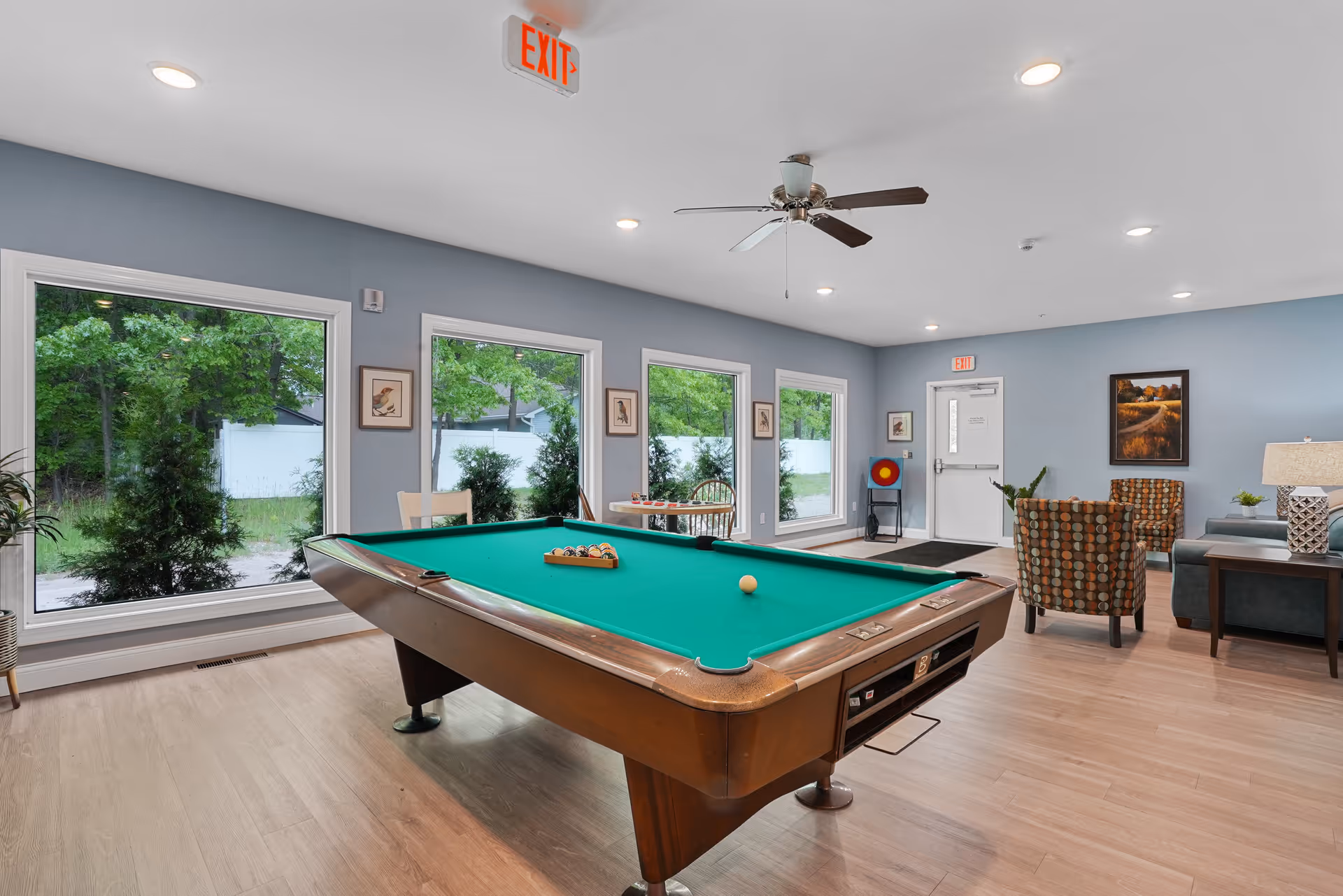 A bright recreational room with a pool table in the center, surrounded by large windows showing green trees outside. The room has light wood flooring, blue walls, a ceiling fan, and recessed lighting. There are two patterned armchairs, a small table with a lamp, and a door with an exit sign above it.