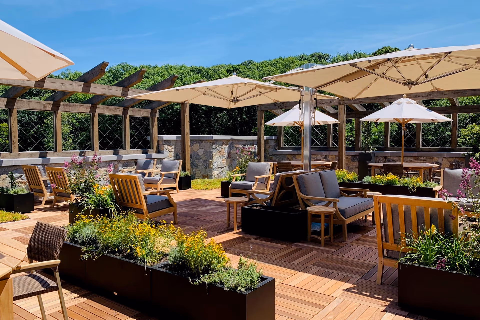 Outdoor patio area with wooden deck flooring, multiple seating arrangements including cushioned chairs and wooden benches, large white umbrellas providing shade, and planters with green and flowering plants. A wooden pergola structure with lattice panels surrounds the area, with trees and a clear blue sky in the background.