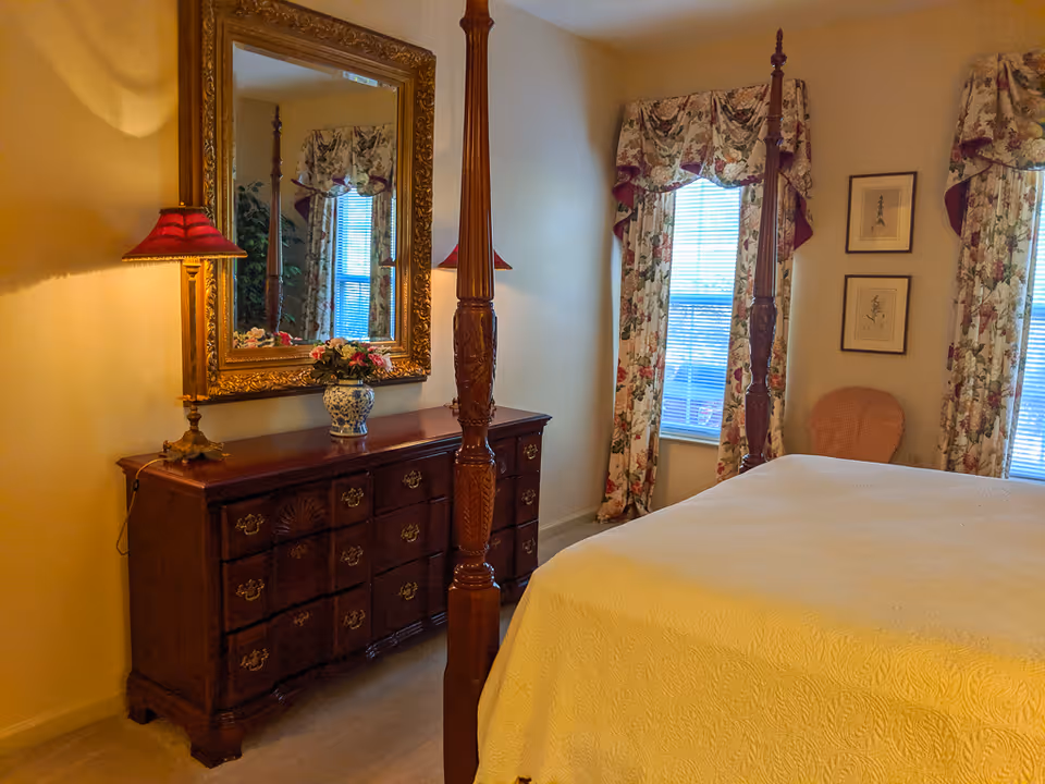 A cozy bedroom featuring a wooden four-poster bed with a cream-colored bedspread. There is a wooden dresser with ornate drawer handles, topped with a decorative vase of flowers and a red lampshade. A large, ornate gold-framed mirror hangs above the dresser. The room has two windows with floral curtains and a wicker chair in the corner. Two framed pictures hang on the wall between the windows.