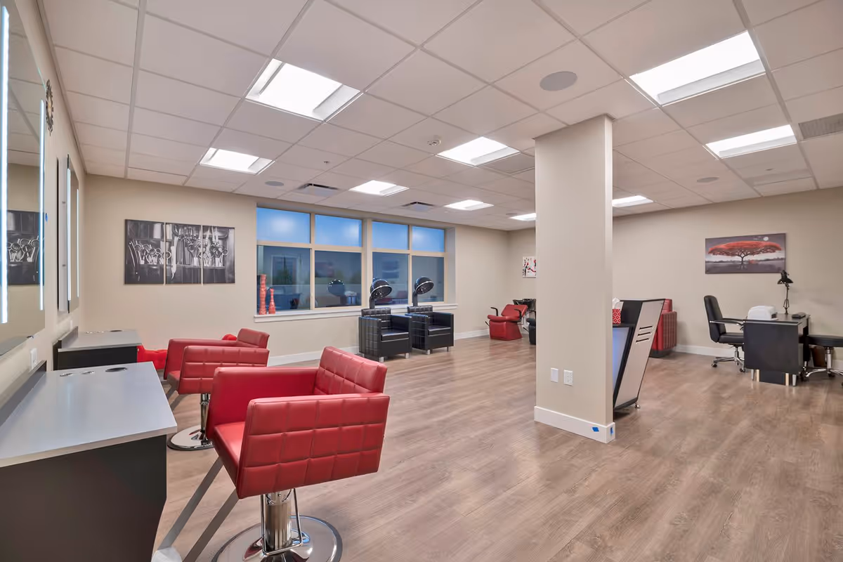 Interior view of a senior living facility's salon area with red salon chairs, black hair drying chairs, a manicure station with a black chair, and modern decor including wall art and large windows.