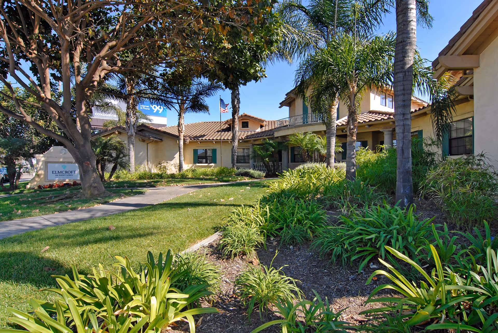 Exterior view of a senior living facility with a well-maintained garden, palm trees, and a walkway leading to the entrance. The building has a tiled roof and beige walls with green window shutters. An American flag is visible near the entrance.