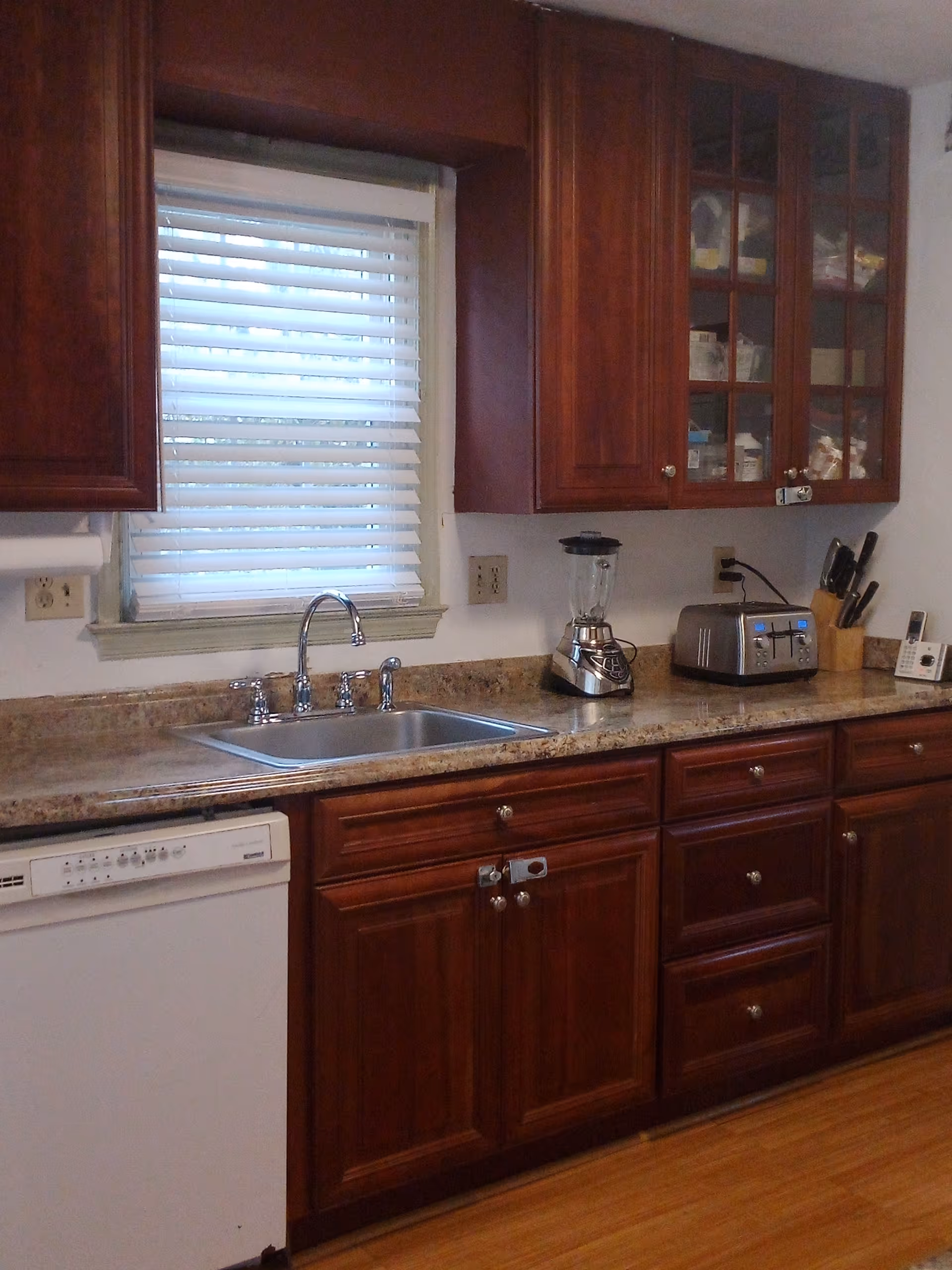A kitchen interior featuring wooden cabinets with silver knobs, a countertop with a sink, a blender, a toaster, a knife block, and a dishwasher. There is a window with white blinds above the sink.