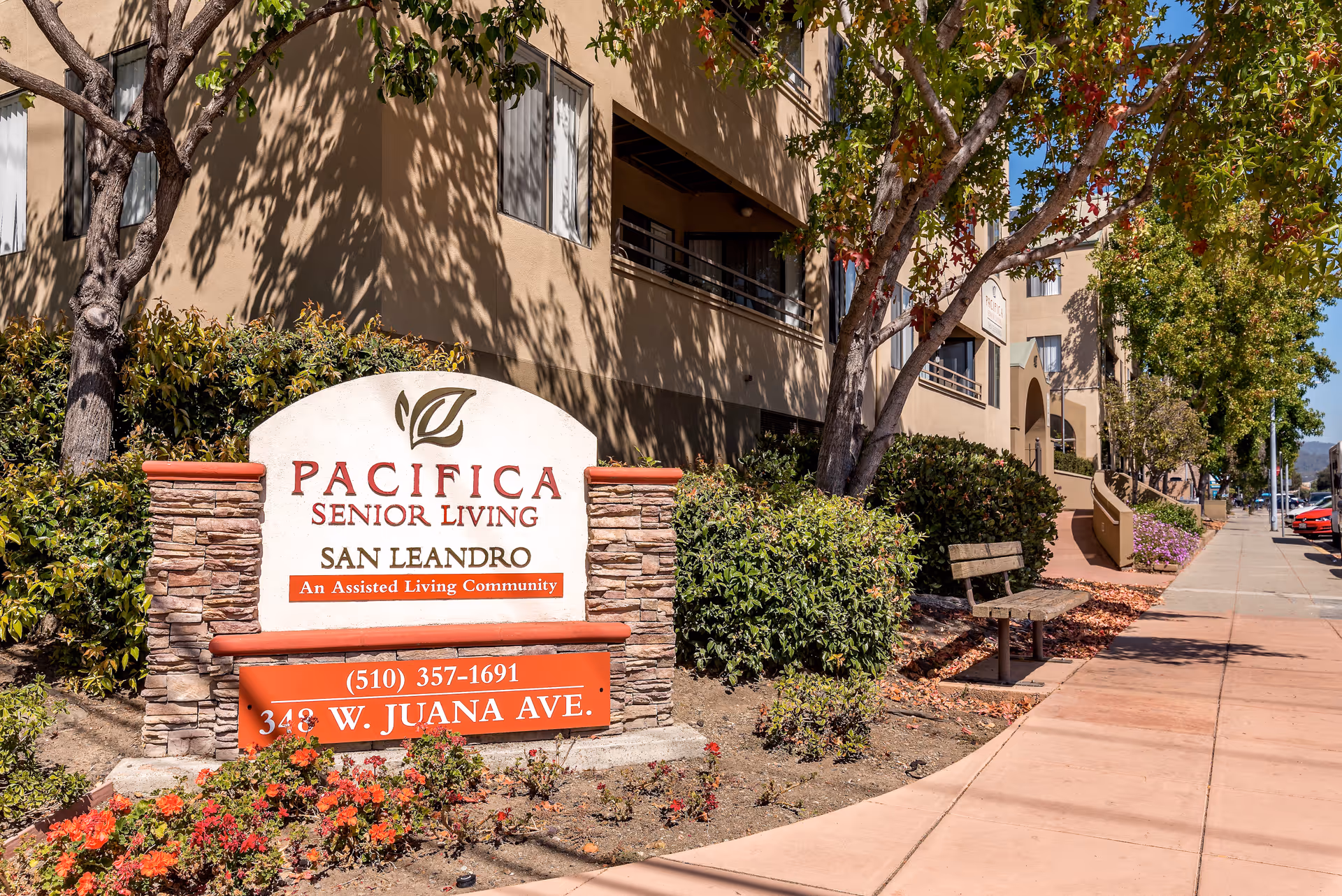 Outdoor view of the entrance sign for Pacifica Senior Living San Leandro, an assisted living community, with the building and sidewalk visible in the background along with trees and bushes.