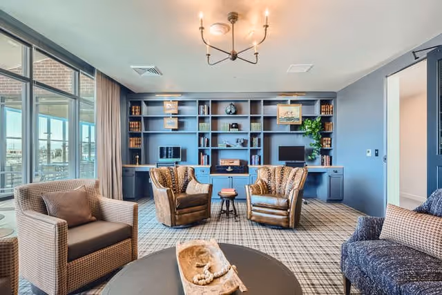 A cozy living room area with a large blue built-in bookshelf filled with books and decorative items. The room features two brown leather armchairs, a woven armchair, and a blue patterned armchair arranged around a round coffee table with a decorative wooden tray. Large windows with curtains allow natural light to fill the space, and a modern chandelier hangs from the ceiling.