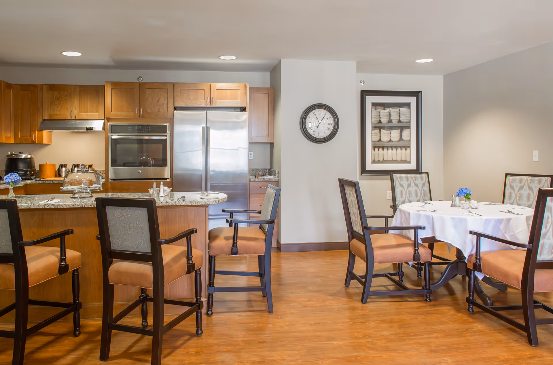 A well-lit dining area and kitchen in a senior living facility. The kitchen features wooden cabinets, a stainless steel refrigerator, an oven, and a granite countertop with bar stools. The dining area has a round table covered with a white tablecloth, surrounded by cushioned chairs. A wall clock and framed artwork are visible on the wall.