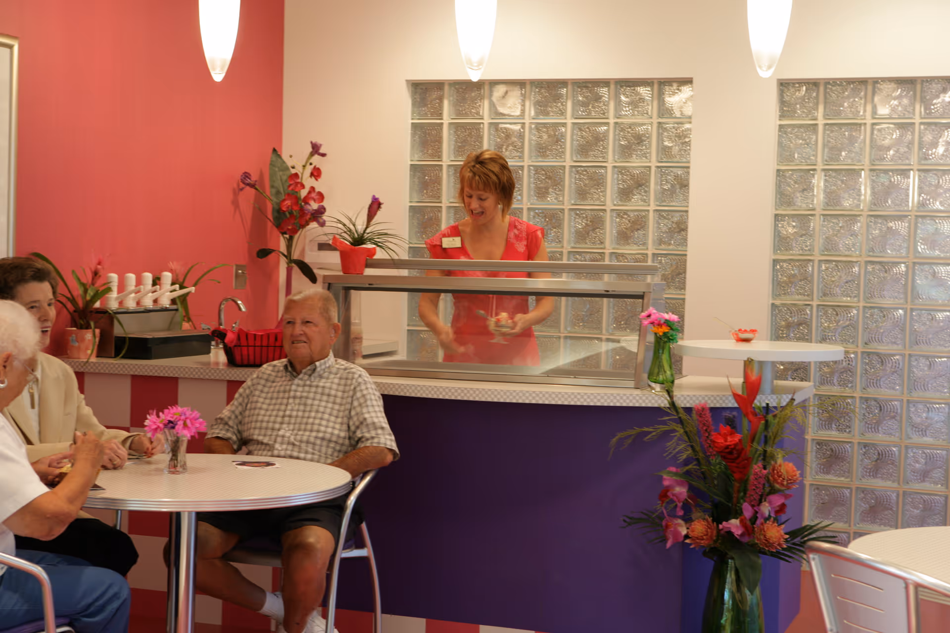 Residents sit at round tables in a colorful dining area while a staff member serves behind a service counter.
