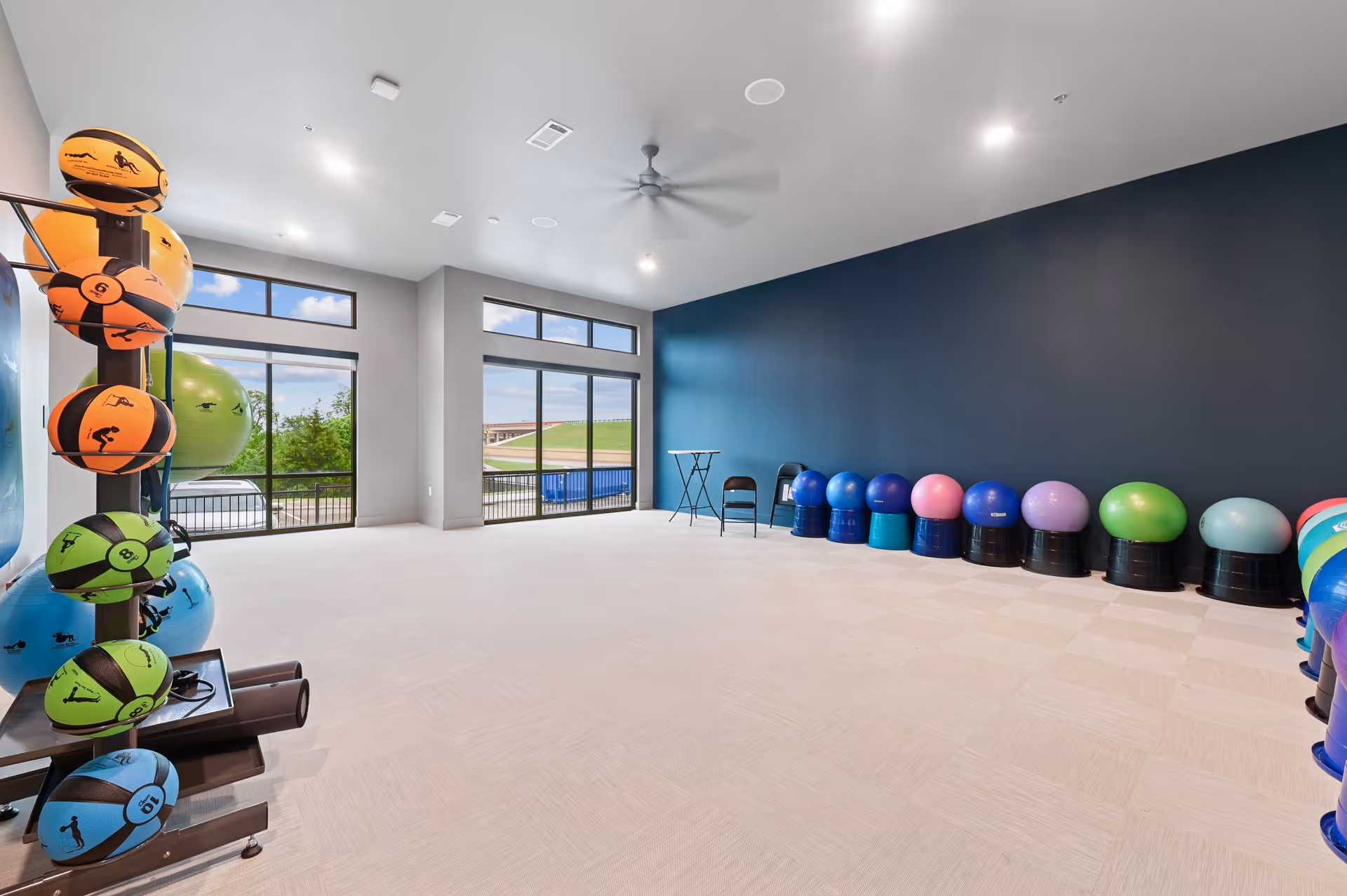 A spacious exercise room with large windows letting in natural light. The room has a rack of colorful medicine balls on the left and a row of variously colored exercise balls on black bases lined up against a dark blue wall on the right. There are two black folding chairs and a small table near the back wall. The floor is light-colored and the ceiling has a large ceiling fan.