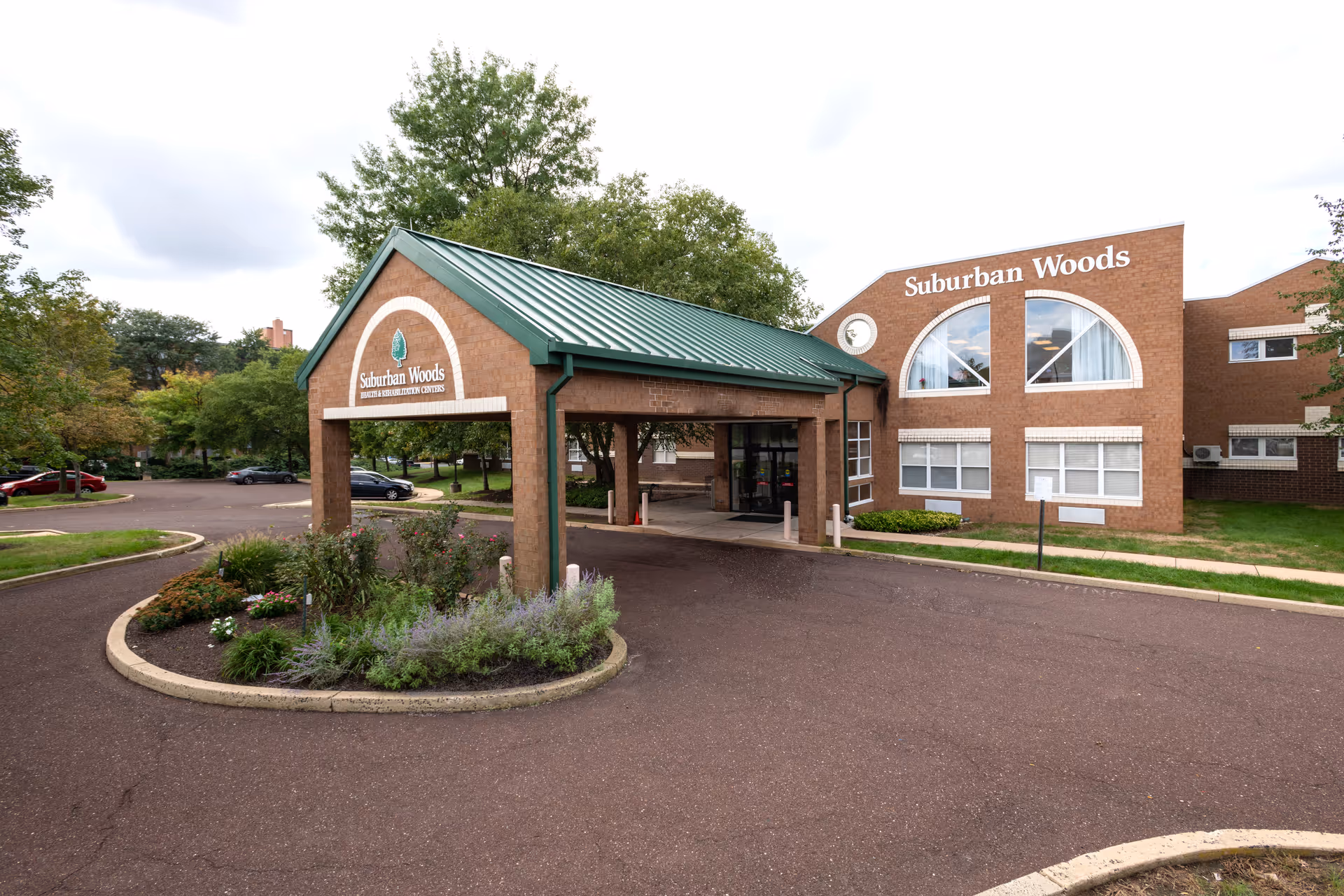 Exterior view of Suburban Woods Health & Rehabilitation Center showing the entrance with a covered driveway, brick building with large windows, and surrounding greenery and parking area.