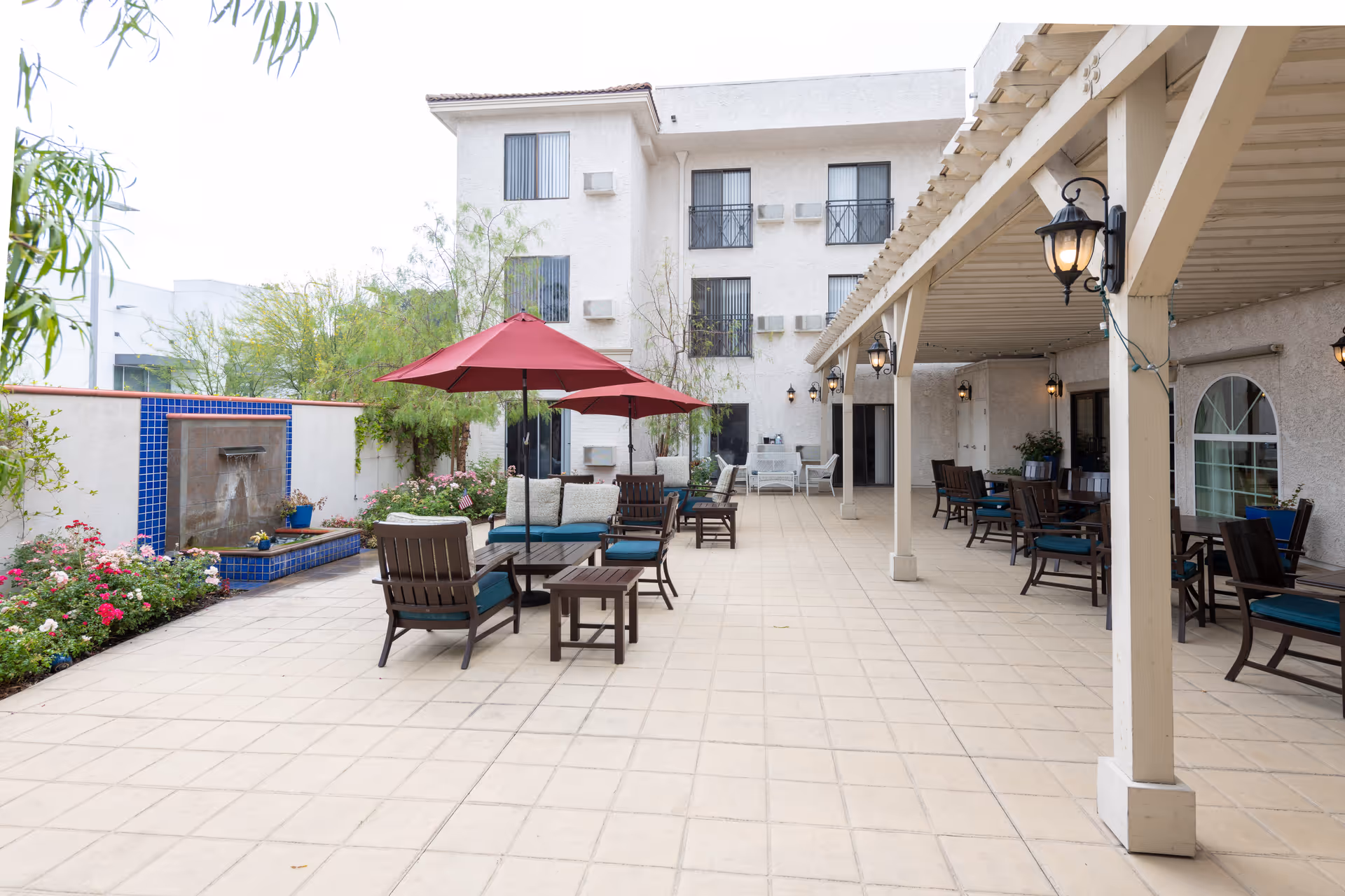 Outdoor patio area at a senior living facility with tiled flooring, several seating arrangements including chairs and tables with red umbrellas, a water fountain with blue tiles on the left, and a covered area with hanging lanterns on the right. The building in the background has multiple windows and air conditioning units.