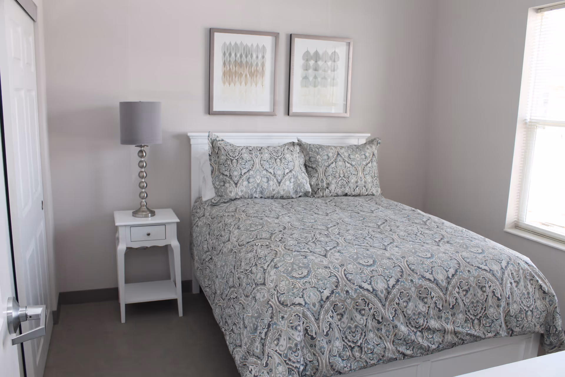 A neatly made bed with a patterned blue and gray comforter and matching pillows in a small bedroom. Next to the bed is a white nightstand with a silver lamp. Two framed abstract artworks hang on the light gray wall above the bed. A window with blinds is on the right side, allowing natural light into the room.