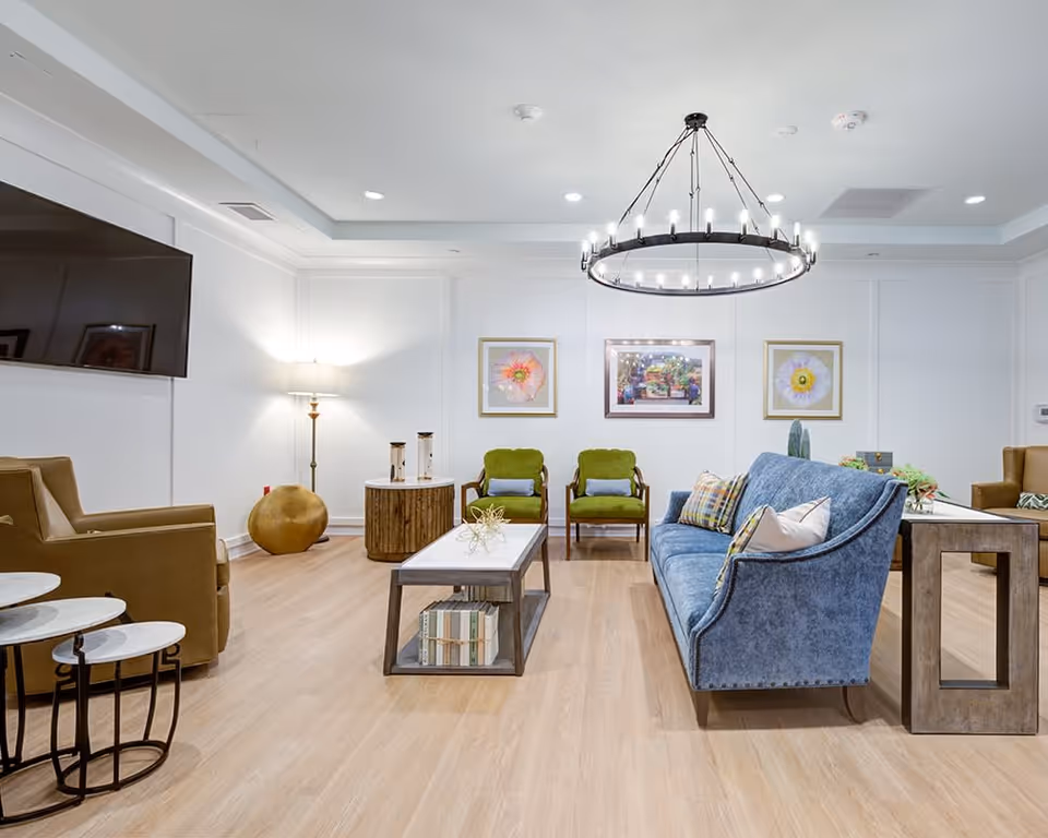 A bright and spacious living room with a blue upholstered sofa, two green armchairs, and two brown armchairs. The room features a modern circular chandelier, a large flat-screen TV mounted on the wall, a wooden coffee table with books underneath, and framed floral artwork on the white walls. The floor is light wood, and there are side tables and decorative items placed around the room.