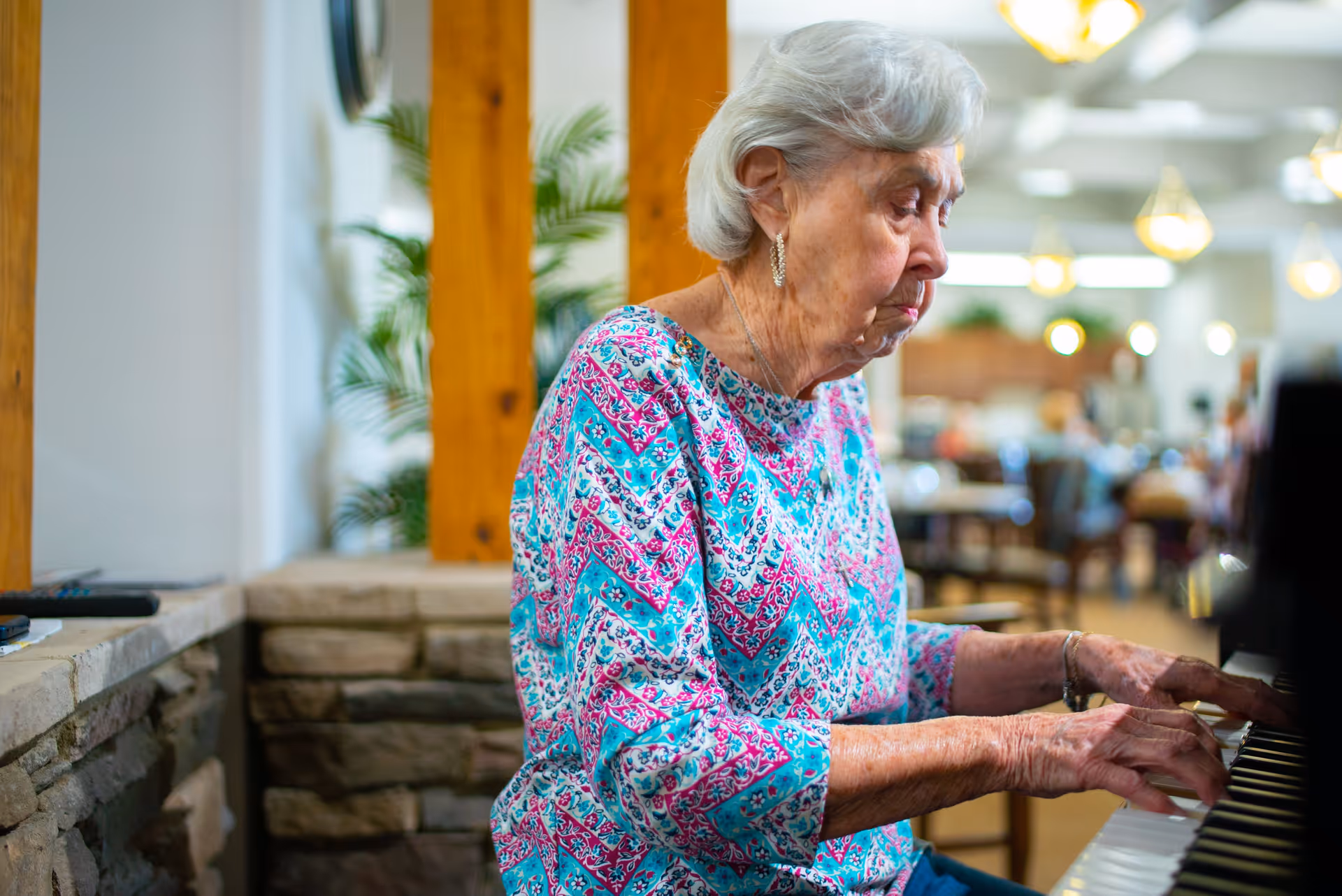 An elderly woman with gray hair wearing a colorful patterned blouse is playing the piano in a well-lit room with wooden beams and stone walls. The background shows a spacious area with tables, chairs, and hanging lights, suggesting a communal or recreational space.