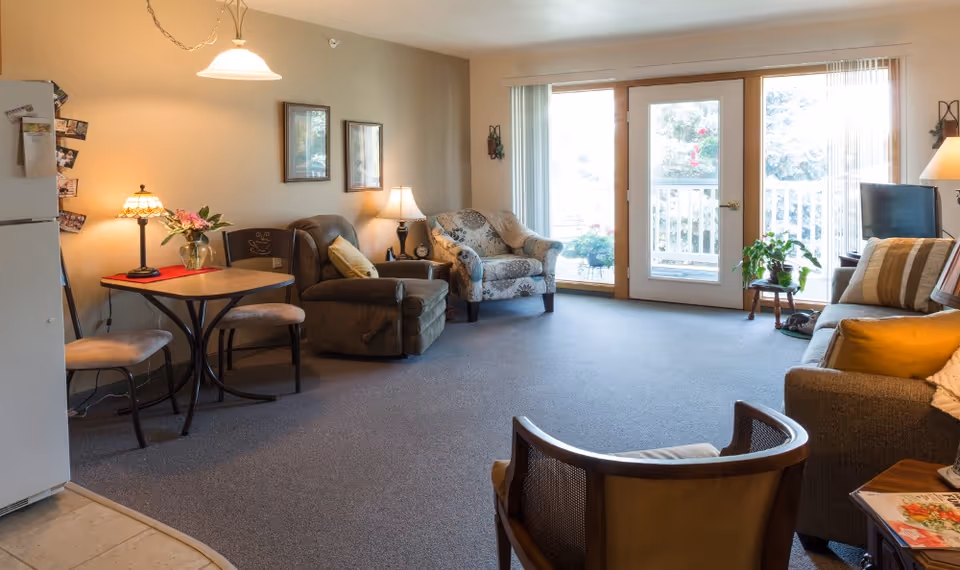 A cozy living room area with a small dining table and two chairs on the left, a brown recliner chair, a patterned armchair, and a sofa with striped pillows. There are two lamps providing warm light, framed pictures on the wall, and large glass doors leading to an outdoor balcony with plants visible outside.