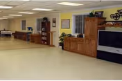 A spacious interior room with a light-colored floor and yellow walls. The room features wooden furniture including a large cabinet with a television, a bookshelf with decorative items, and a bench along the far wall. There are windows with white blinds and ceiling lights illuminating the space.