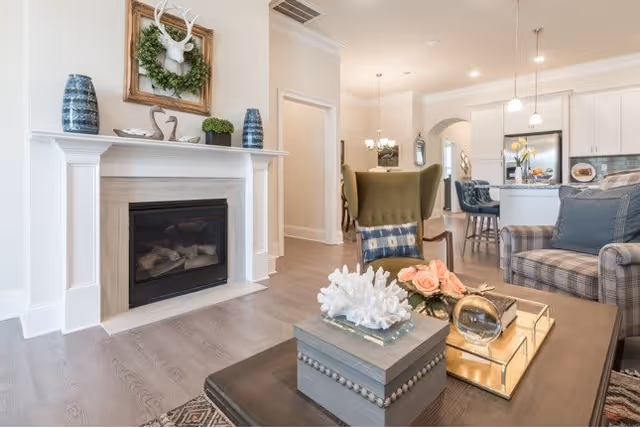 A bright and cozy living room area featuring a fireplace with decorative vases and a framed deer head with a wreath above it. In the foreground, a coffee table holds decorative items including coral, flowers, and a glass orb. The room opens into a dining area with a green wingback chair and a kitchen with white cabinetry and pendant lights.