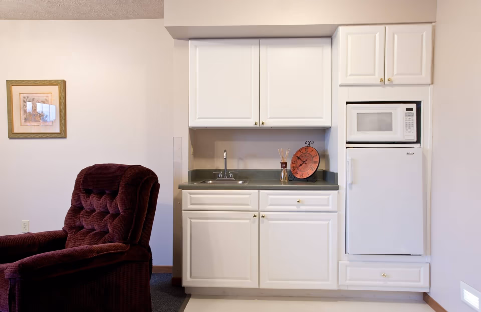 A small kitchenette area with white cabinets, a countertop with a sink, a microwave, and a mini refrigerator. To the left of the kitchenette is a plush maroon recliner chair and a framed picture hanging on the wall.