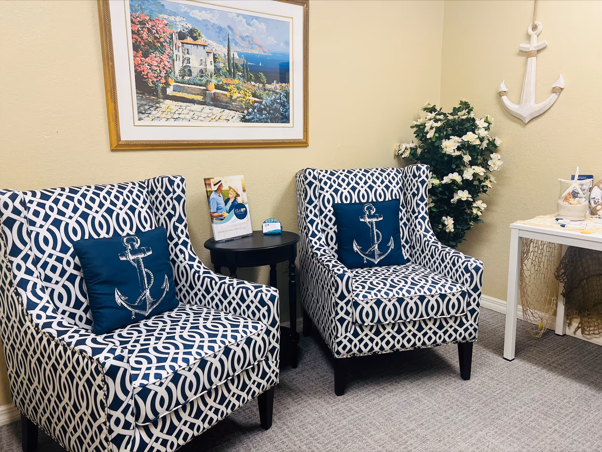 Two patterned armchairs with navy blue and white geometric designs, each with a navy blue pillow featuring a white anchor. A small round black table is placed between the chairs with a brochure and a small item on it. A framed painting of a coastal scene hangs on the beige wall above the chairs. To the right, there is a white table with decorative items and a green plant with white flowers in the corner. A white anchor decoration is mounted on the wall above the plant.
