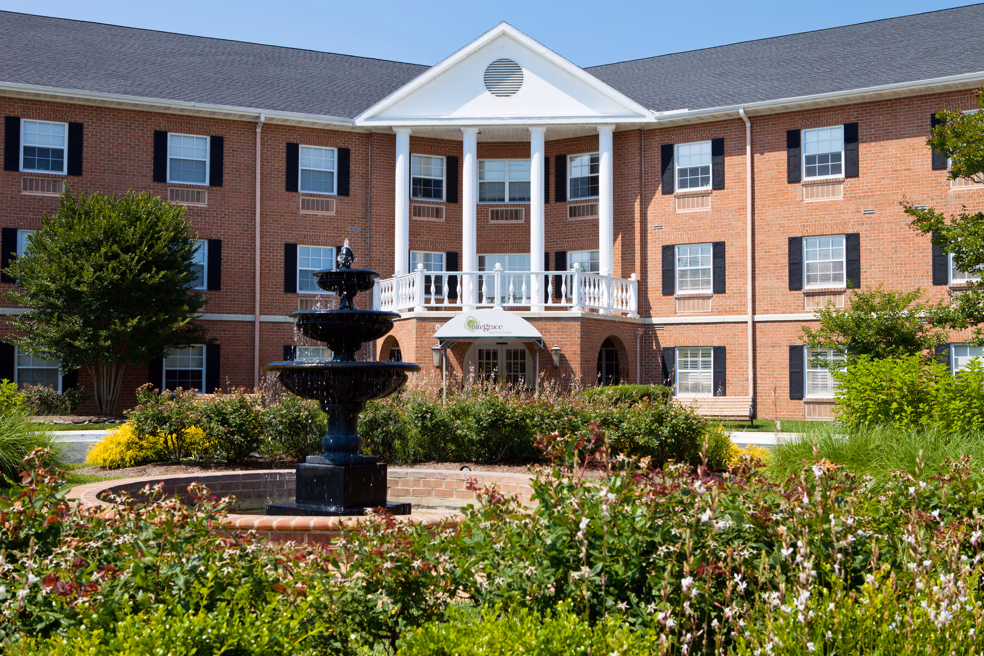Brick Bayleigh Chase senior living building with white columns, balcony, and central fountain in landscaped grounds.