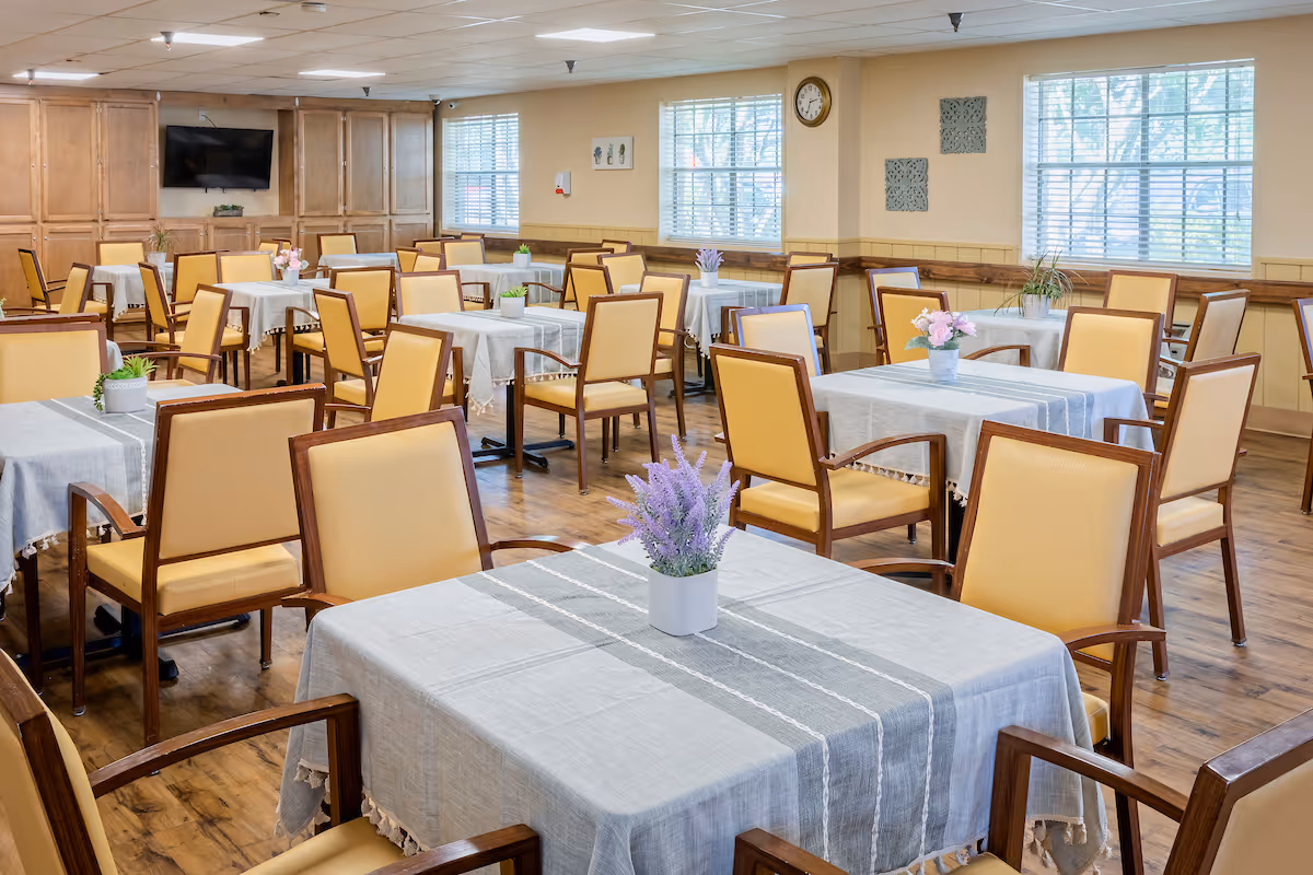 A bright dining room with multiple tables covered with light gray tablecloths and decorated with small flower pots. The room has wooden floors, yellow cushioned chairs with wooden frames, large windows with blinds, and a wall-mounted TV.