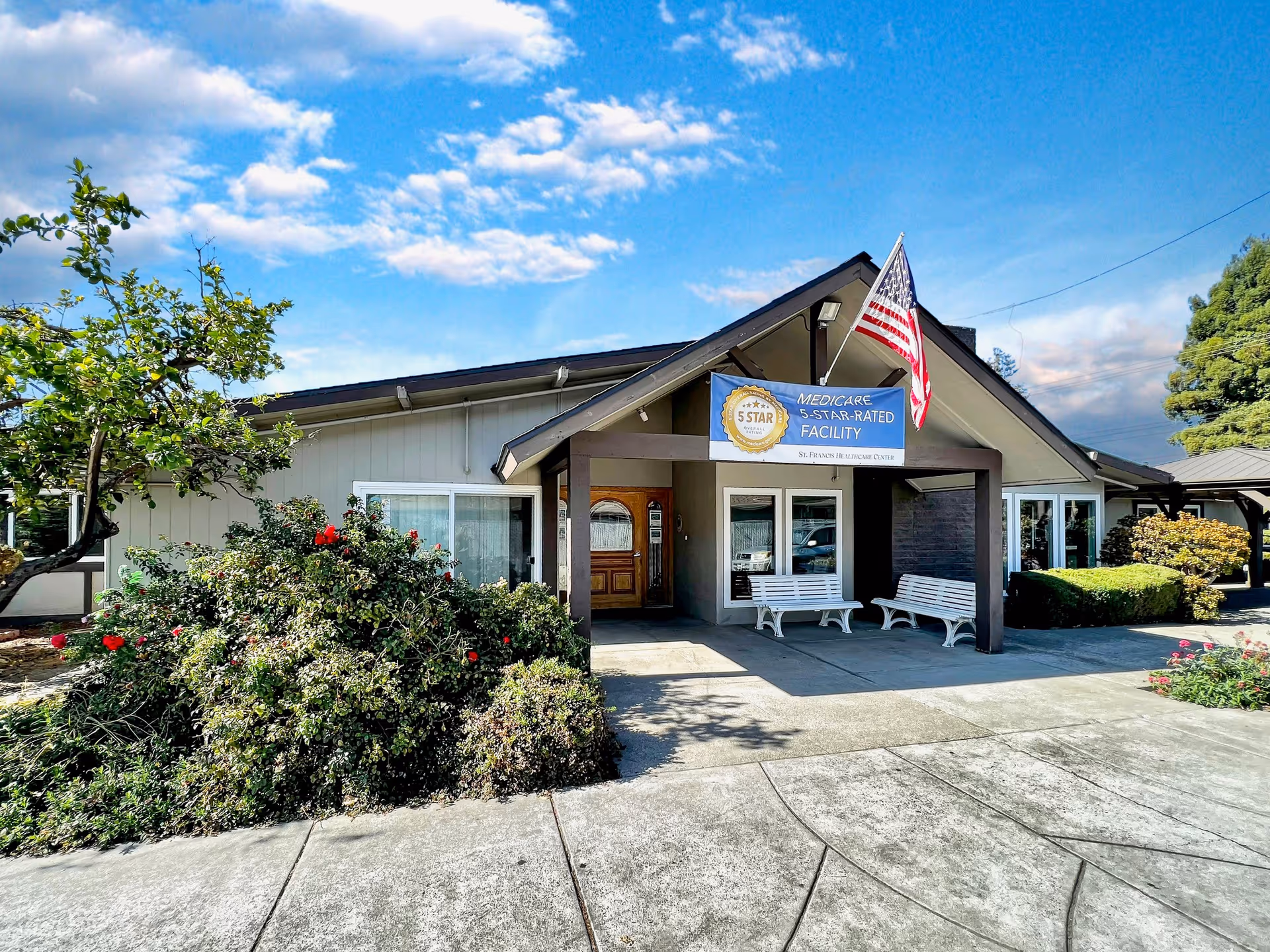 Exterior view of St. Francis Healthcare Center building with a covered entrance, two white benches, an American flag, and a sign indicating it is a Medicare 5-star-rated facility. The building is surrounded by greenery and bushes under a partly cloudy blue sky.
