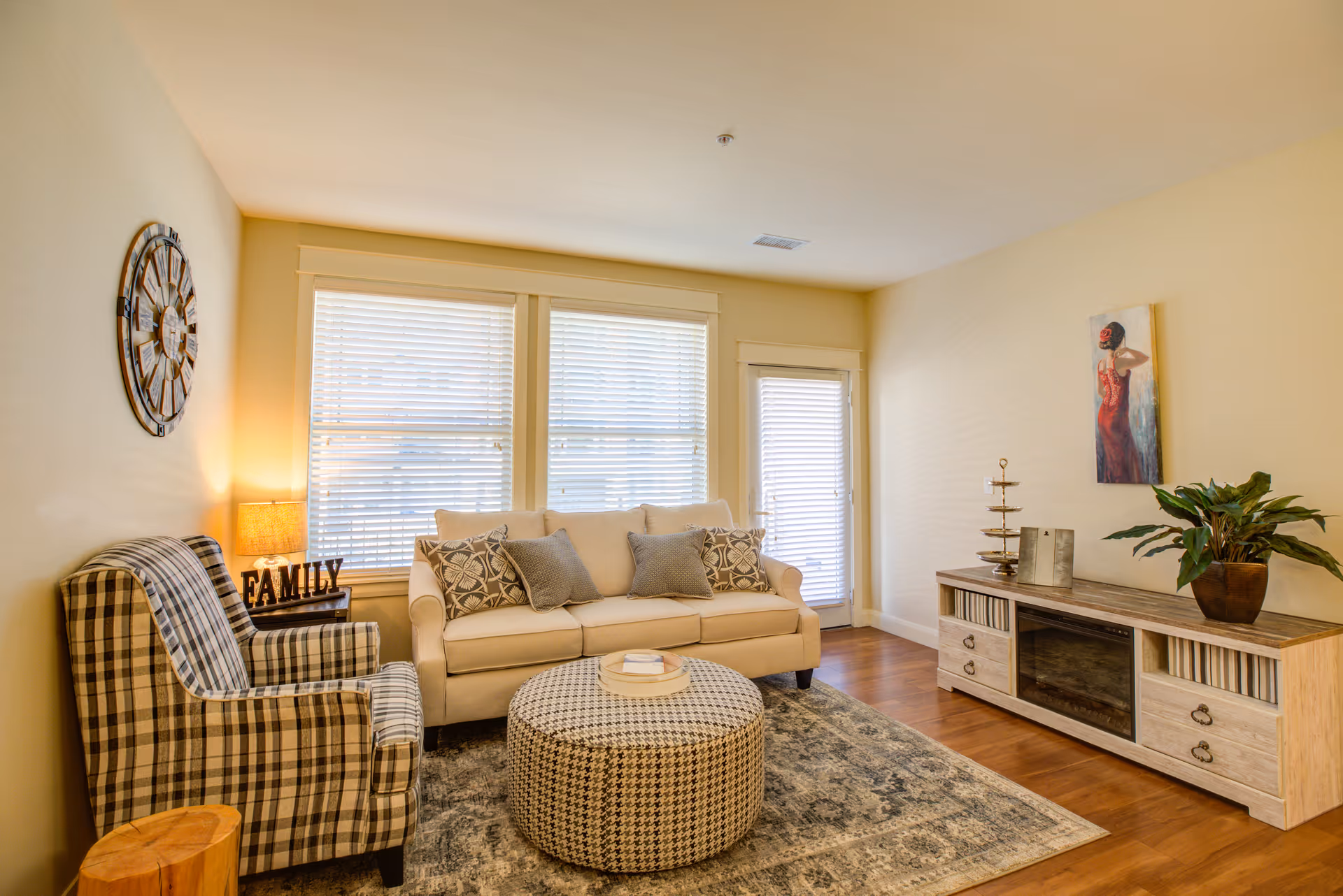 A cozy living room with a beige sofa adorned with patterned cushions, a plaid armchair, a round patterned ottoman, and a wooden entertainment unit with a plant and decorative items. The room has wooden flooring, a large wall clock, a lamp on a side table, and two windows with blinds letting in natural light.