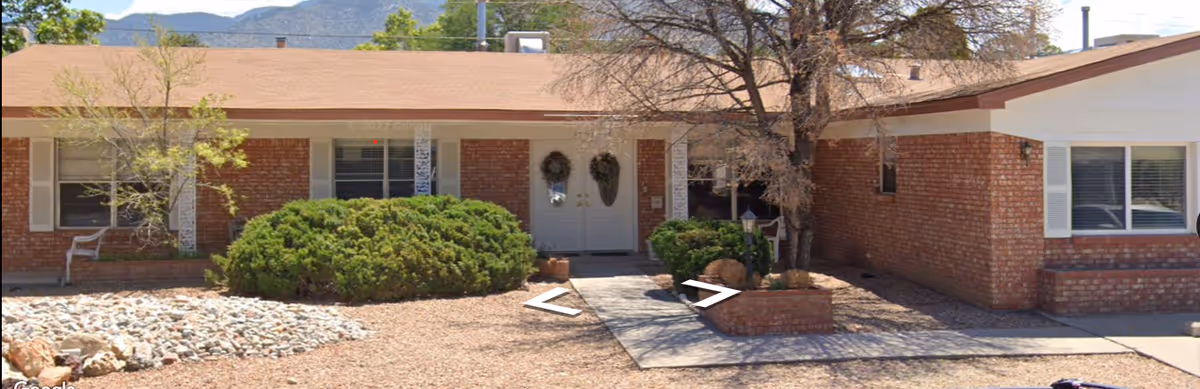 Exterior view of a single-story brick building with a tan roof, featuring a central entrance with double doors decorated with wreaths. There are bushes and small trees in the front yard, with a pathway leading to the entrance. The sky is clear and mountains are visible in the background.