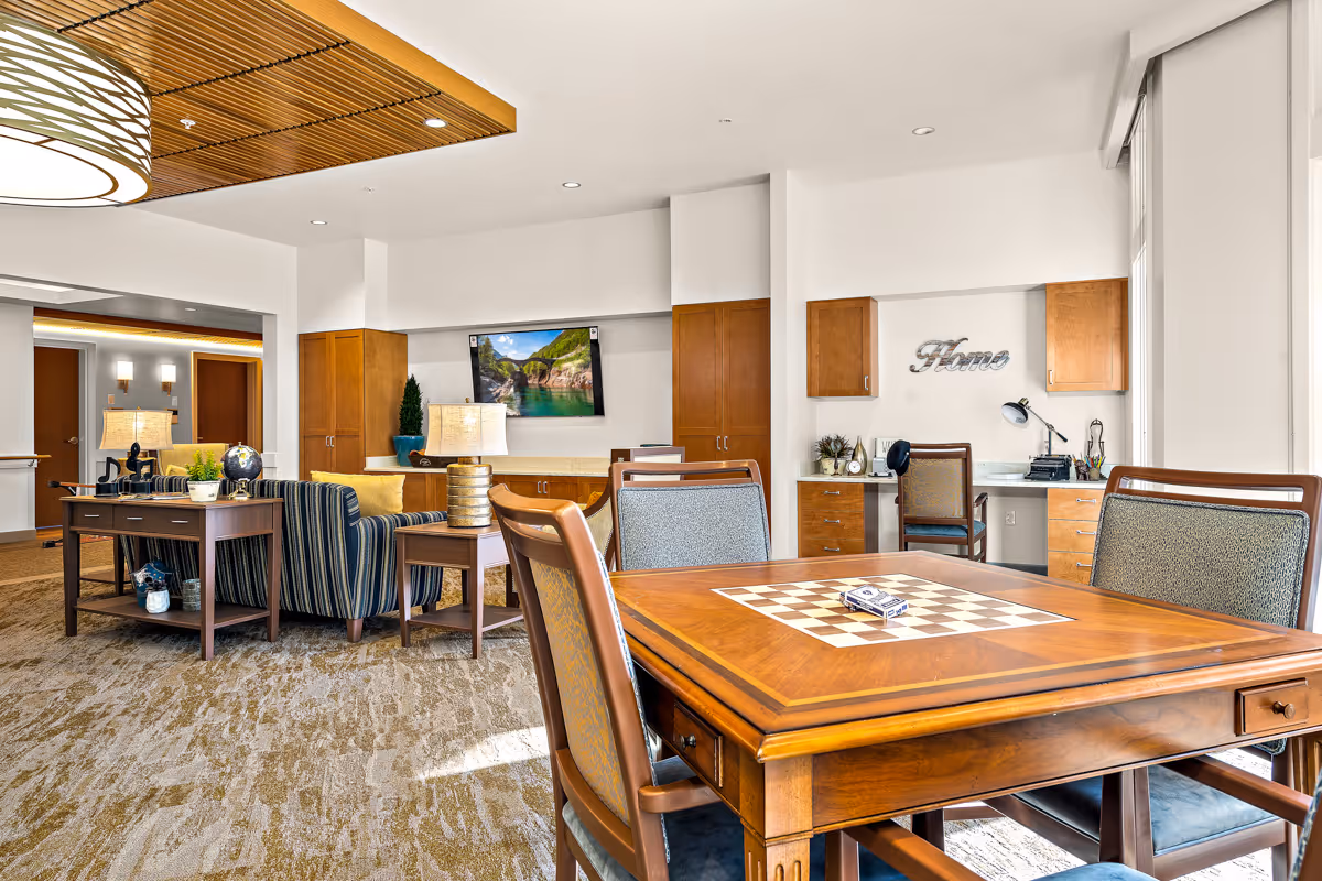 A bright and spacious common area in a senior living facility featuring a wooden table with a built-in chessboard and four chairs. In the background, there is a seating area with striped armchairs, a wooden console table with lamps and decorative items, and a wall-mounted TV displaying a nature scene. The room also includes a small desk area with a chair, desk lamp, and decorative plants, with the word 'Home' displayed on the wall. The space has carpeted flooring, wooden cabinetry, and large windows allowing natural light to fill the room.