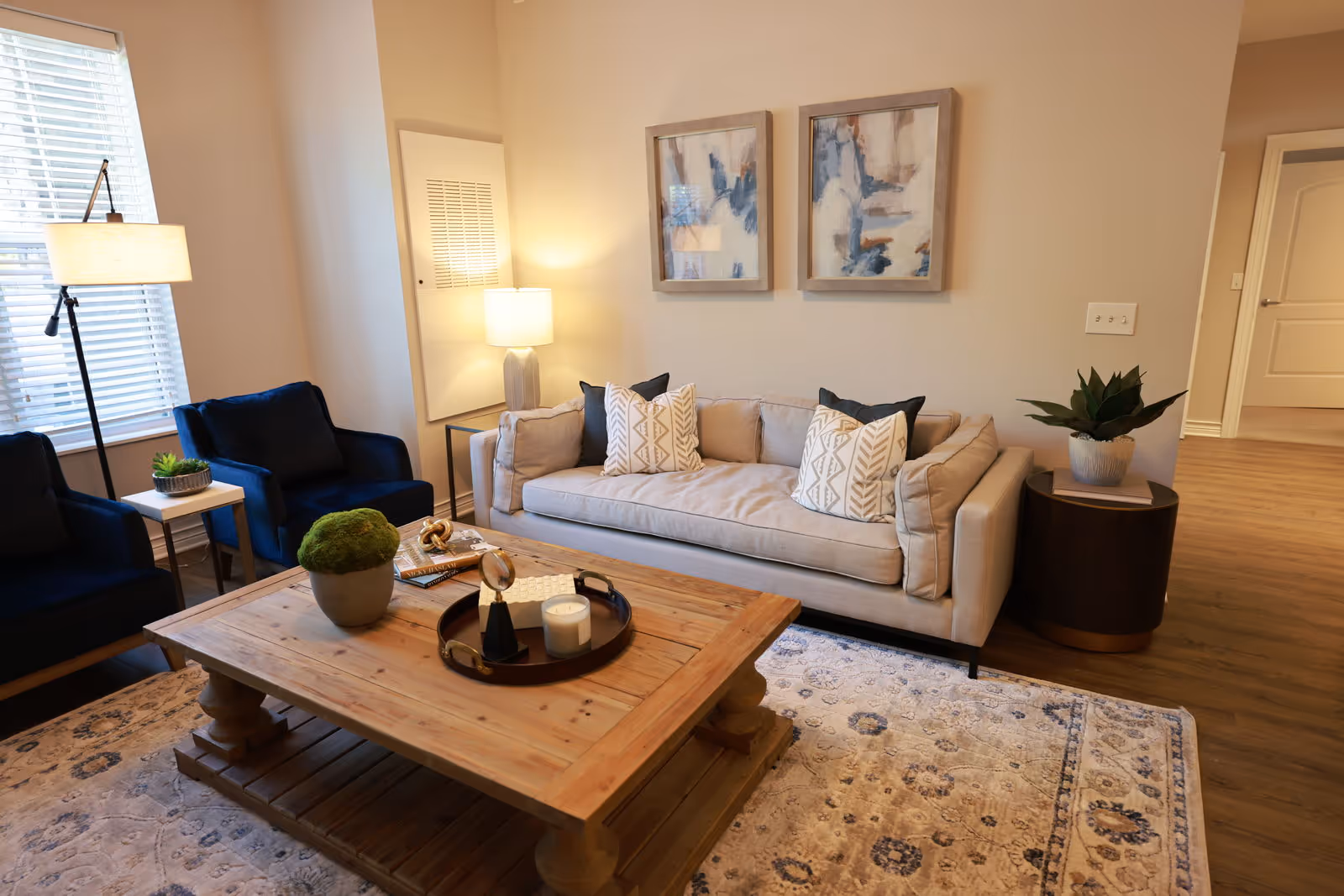 A bright furnished living room with a beige sofa, two blue armchairs, a wooden coffee table, and wall artwork.