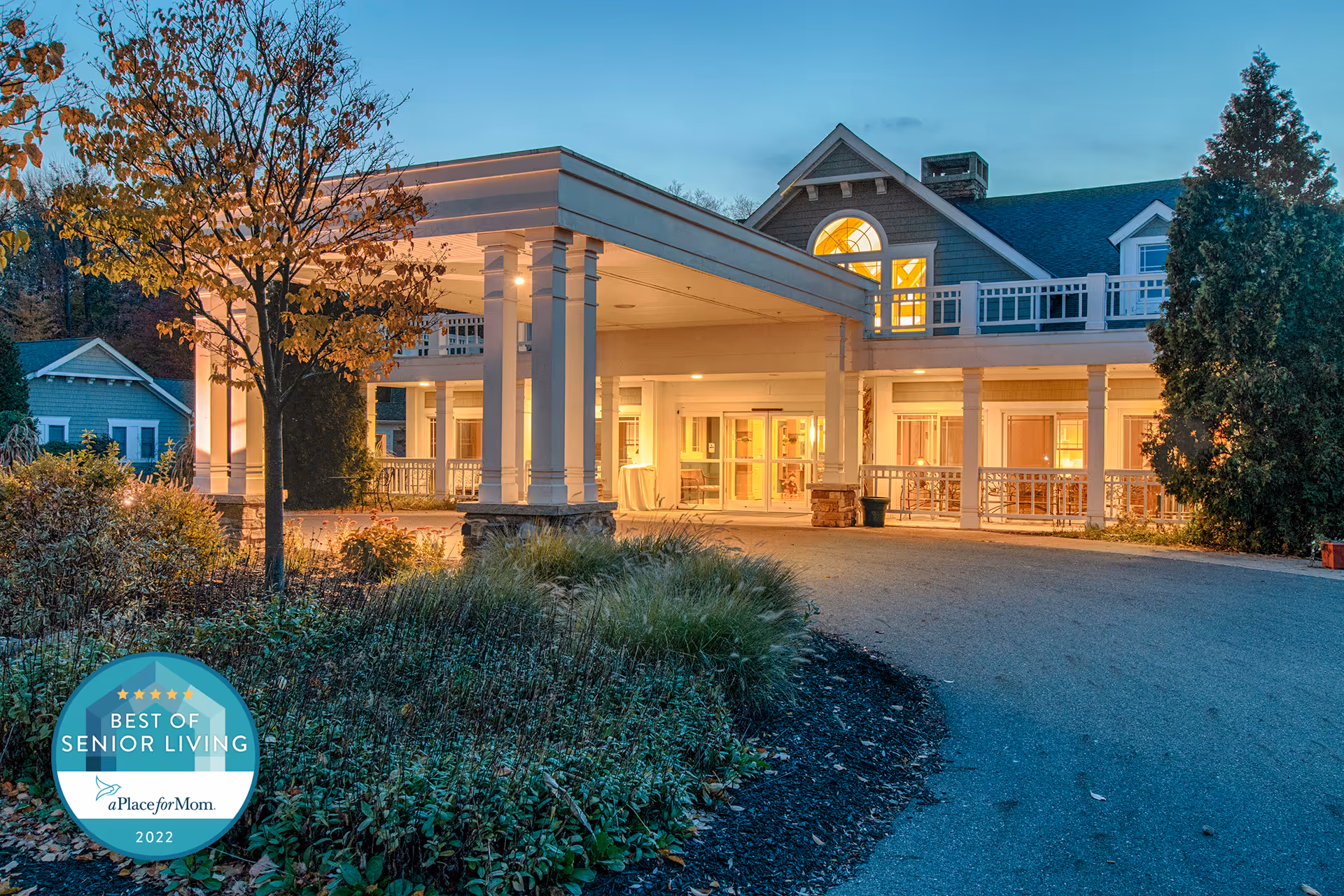 Exterior view of a senior living facility at dusk with warm lights glowing from inside. The building has a covered entrance supported by columns, surrounded by trees and landscaped greenery. A circular driveway leads up to the entrance.