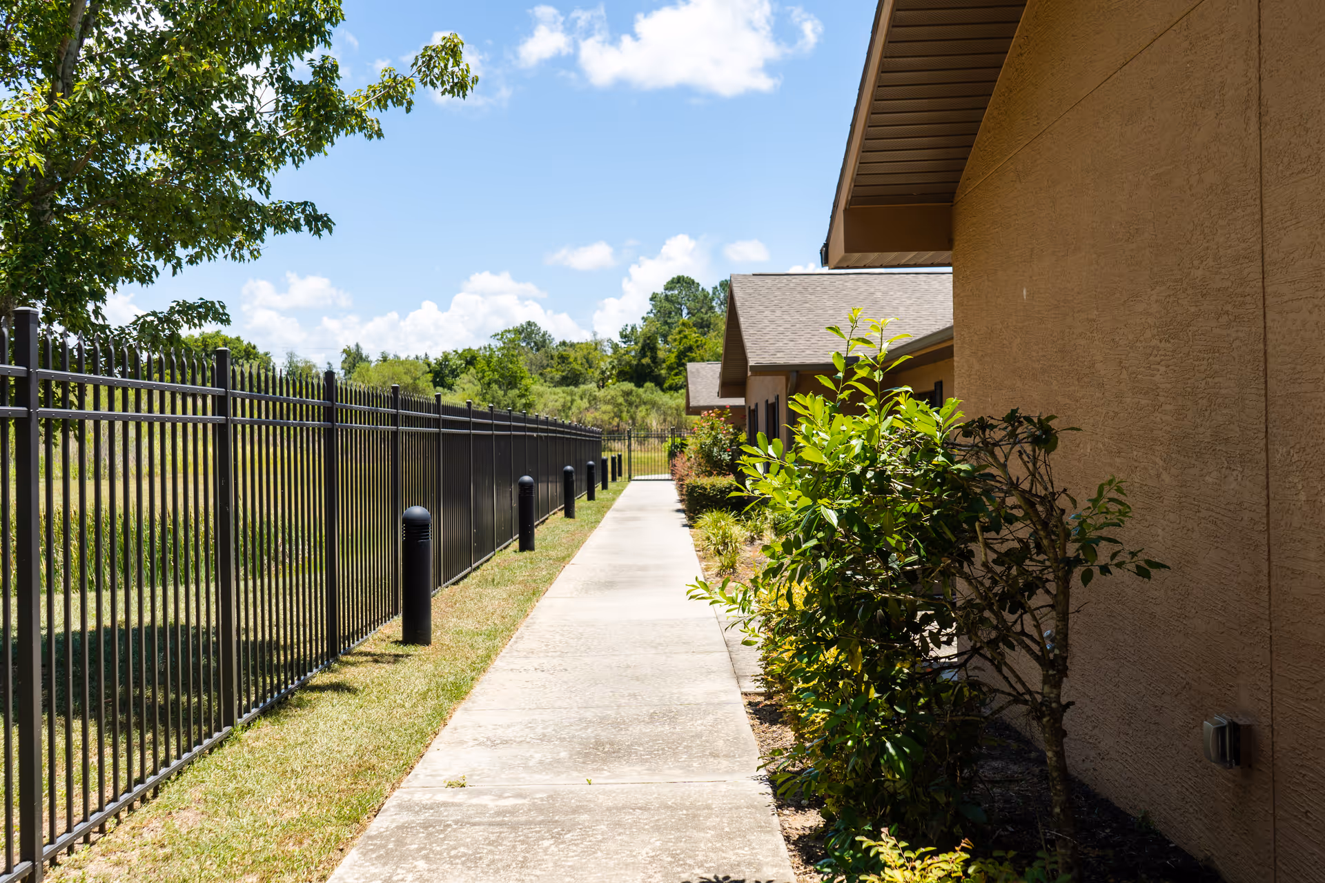 A sunny outdoor walkway alongside a beige building with a black metal fence on the left and green bushes on the right under a blue sky with some clouds.