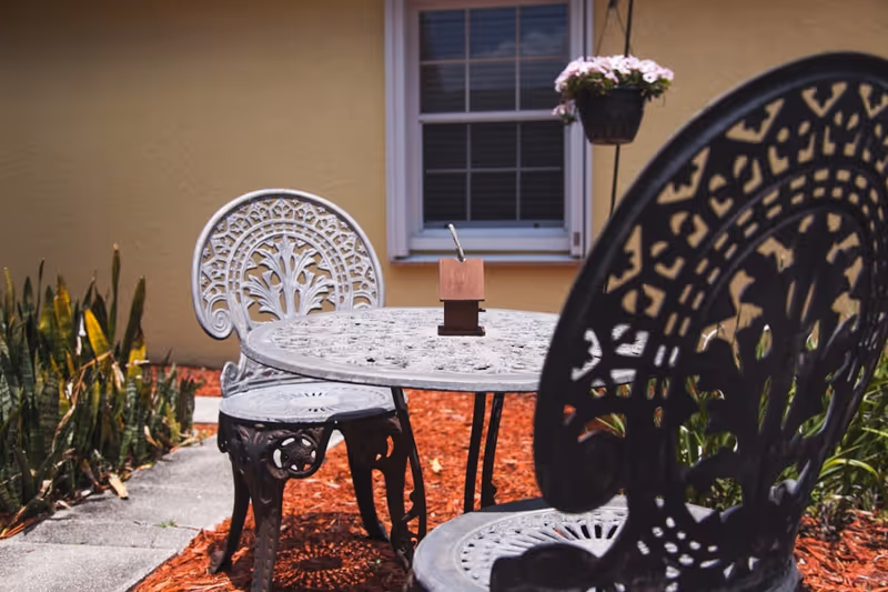 Outdoor patio area with a decorative metal table and two matching chairs on a concrete path surrounded by red mulch and plants. A hanging flower pot is visible in front of a window on a beige building wall.
