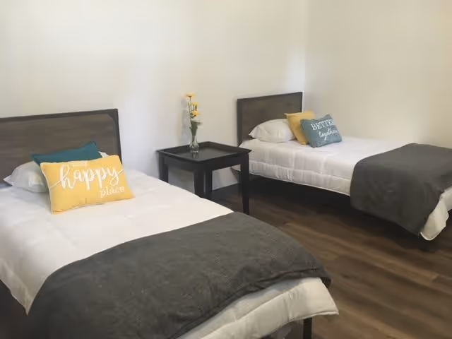 A simple bedroom with two single beds, each with white bedding, gray blankets, and decorative pillows. One pillow says 'happy place' and the other says 'better together'. A small black nightstand with a vase of yellow flowers is placed between the beds. The room has wooden flooring and plain white walls.