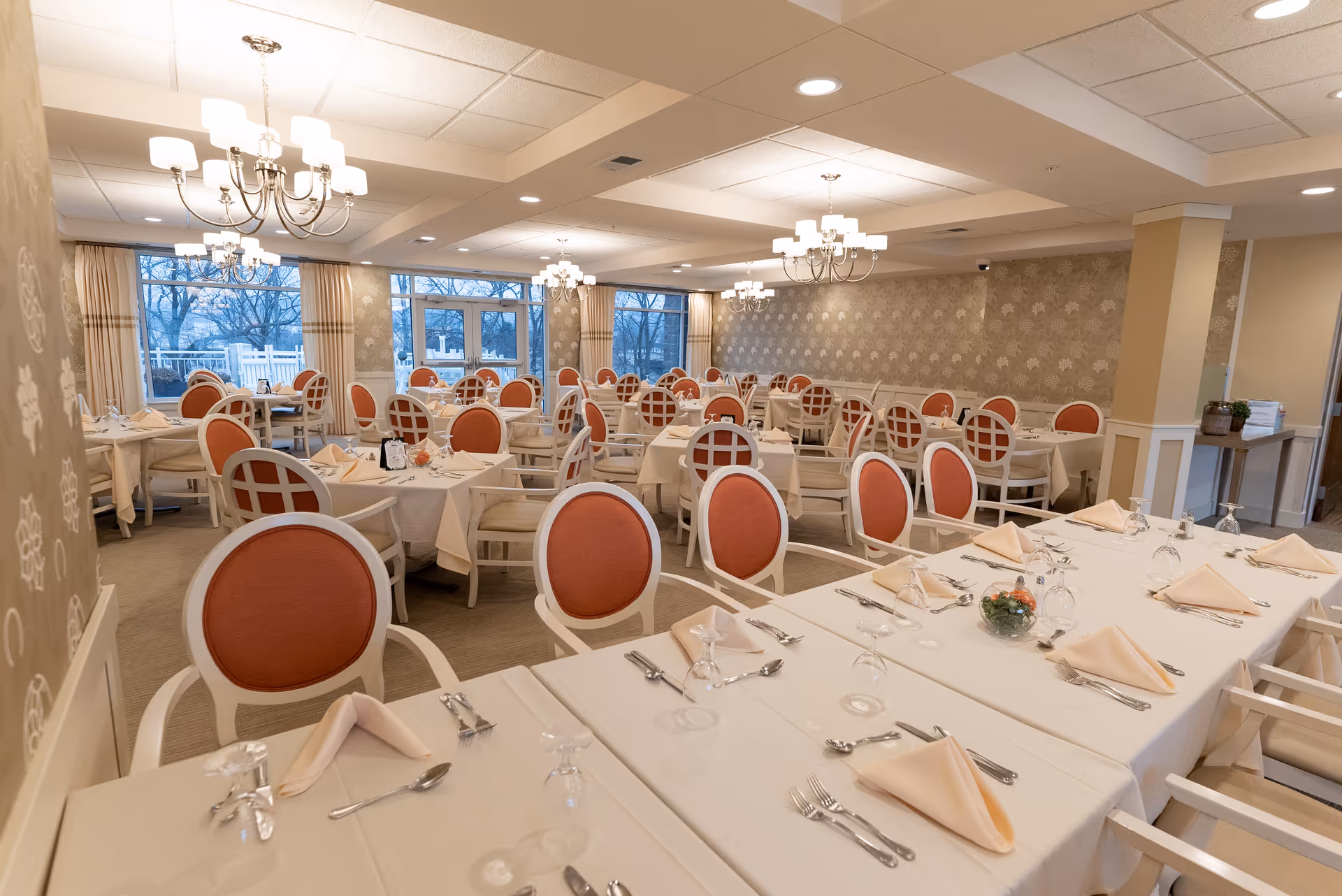A spacious dining room with multiple tables covered in white tablecloths, each set with beige napkins, silverware, and upside-down glasses. The chairs have white frames with orange cushioned seats and backs. The room is well-lit with chandeliers hanging from the ceiling and large windows along one wall showing a view of trees outside.