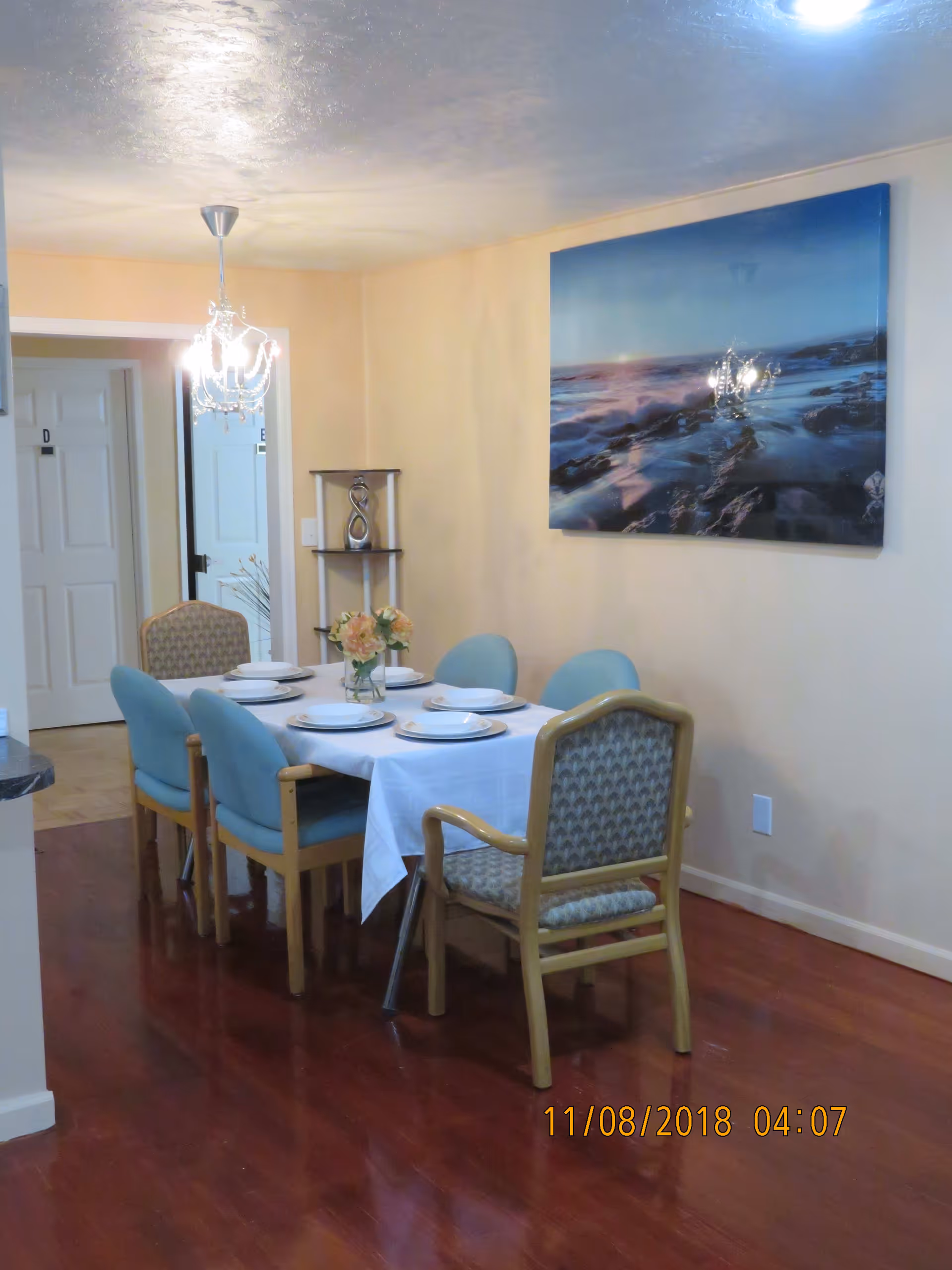 A dining room with a rectangular table covered with a white tablecloth set with six place settings. The table is surrounded by six chairs, four with blue cushions and two with patterned upholstery. A small vase with flowers is centered on the table. A chandelier hangs from the ceiling above the table. On the wall to the right, there is a large framed picture of a rocky shoreline at sunset. In the background, there are two white doors labeled with letters D and E. The floor is wooden with a polished finish.