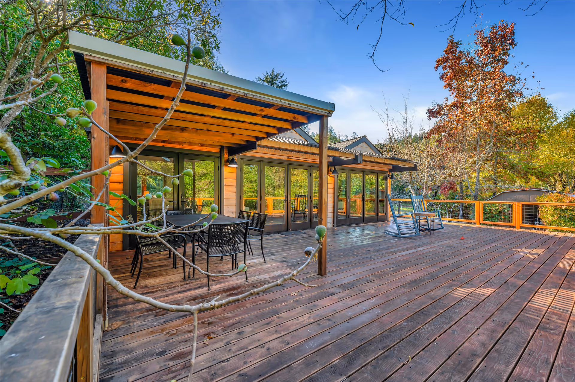 A spacious wooden outdoor deck attached to a building with large glass doors. The deck features a covered seating area with a table and chairs, as well as two rocking chairs. Surrounding the deck are trees with green and autumn-colored leaves under a clear blue sky.