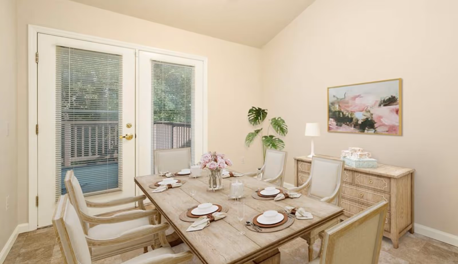 A dining room with a wooden dining table set for six people. The table has plates, napkins, glasses, and a vase with pink flowers. There are six cushioned chairs around the table. Behind the table is a wooden sideboard with a white lamp and decorative boxes on top. A large abstract painting with pink and green hues hangs on the wall above the sideboard. French doors with blinds lead to an outdoor area.