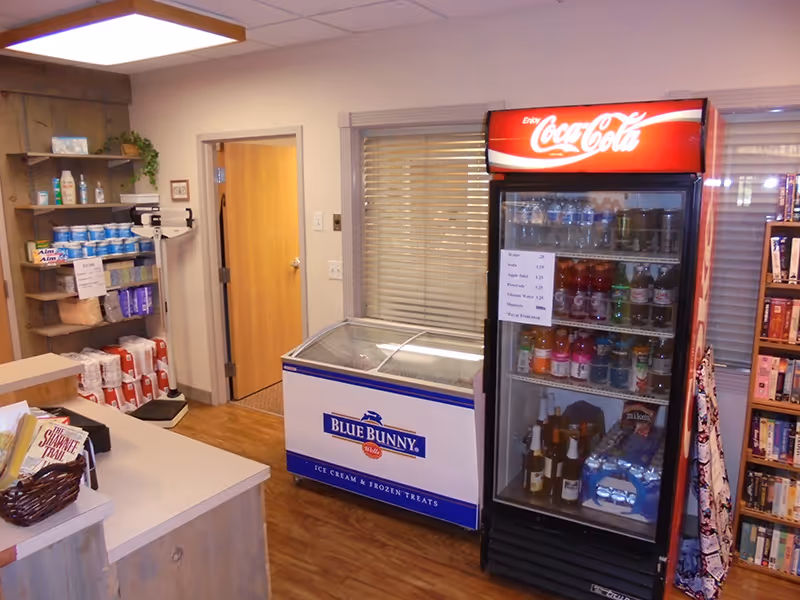 Interior view of a small convenience area in an assisted living community featuring a Coca-Cola beverage refrigerator stocked with drinks, a Blue Bunny ice cream freezer, shelves with various toiletries and paper products, and a counter with a basket of snacks. There is a door and a window with blinds in the background.
