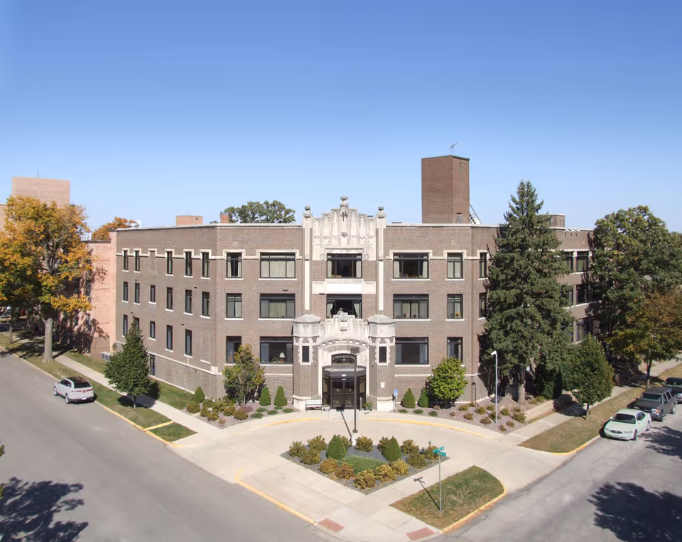 Exterior view of a three-story brick building with a decorative entrance labeled Cornerstone. The building is surrounded by trees and shrubs, with a clear blue sky above. Cars are parked along the streets on either side of the building.